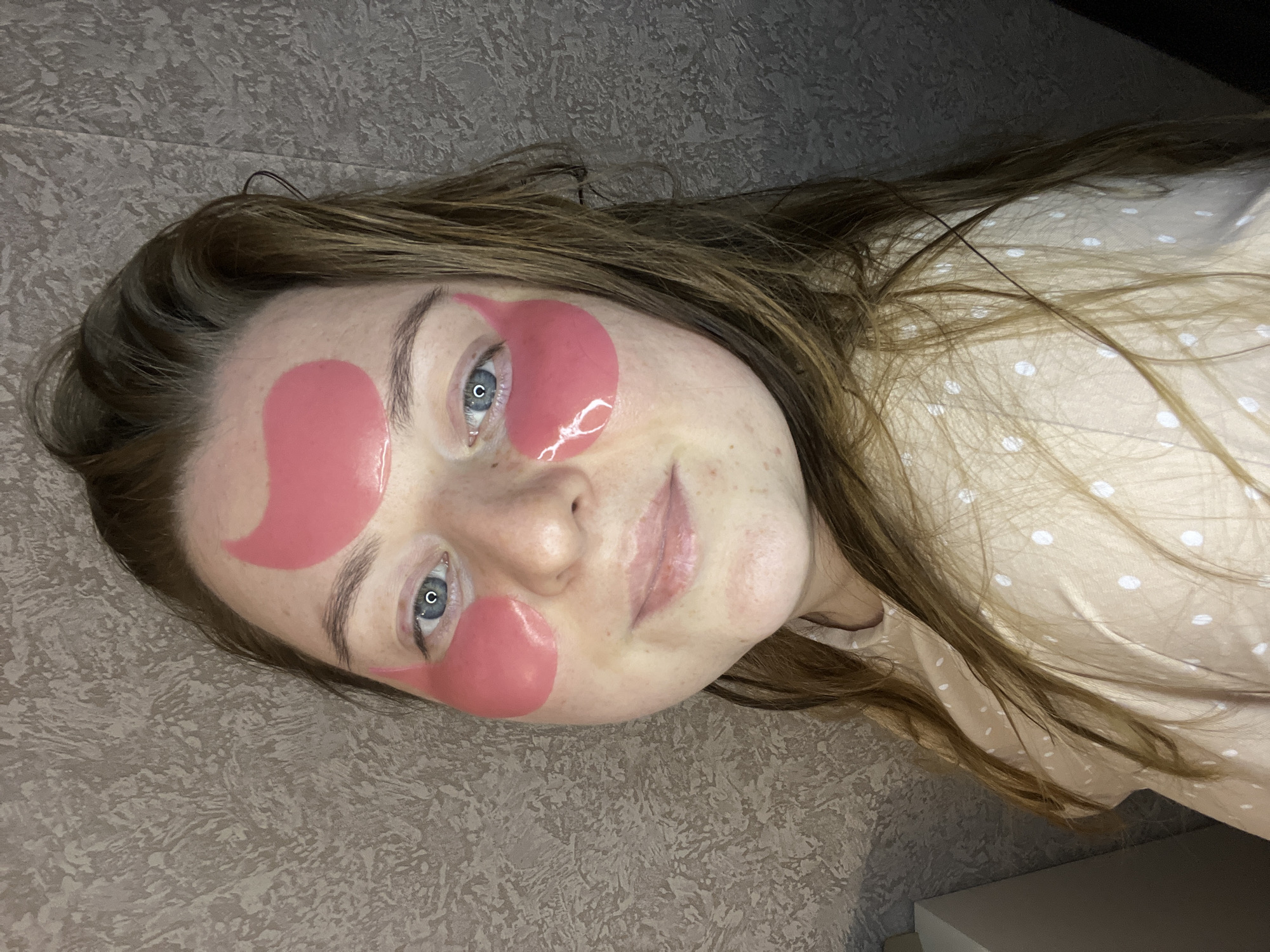 A woman with light brown hair and pink eye patches is lying down on a beige surface. She is wearing a light beige shirt with white polka dots. The background is a textured beige surface.