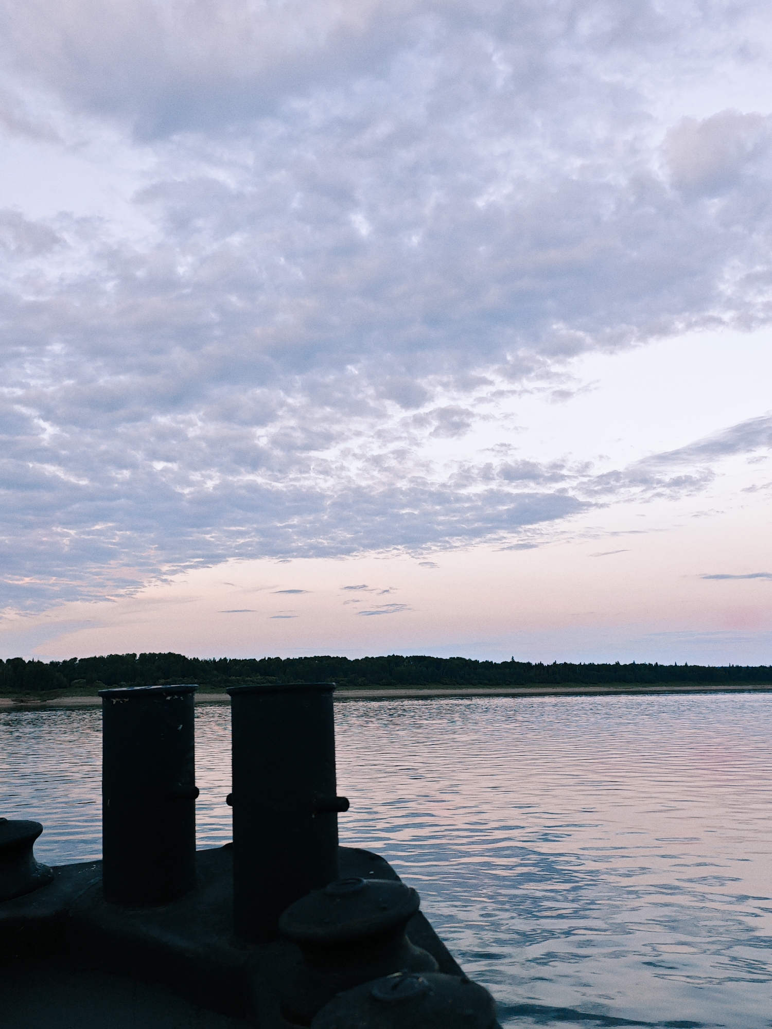 A tranquil scene unfolds at dusk on the river in Nikulino, Russia. The sky is a canvas of soft blues and pinks, adorned with wispy clouds. In the foreground, the dark, silhouetted forms of two bollards rise from the deck of a vessel, anchored in the calm waters. The river reflects the hues of the twilight sky, creating a shimmering surface. Across the water, a lush green forest line marks the distant shore. The overall atmosphere is one of peaceful stillness, characteristic of a serene evening by the river.