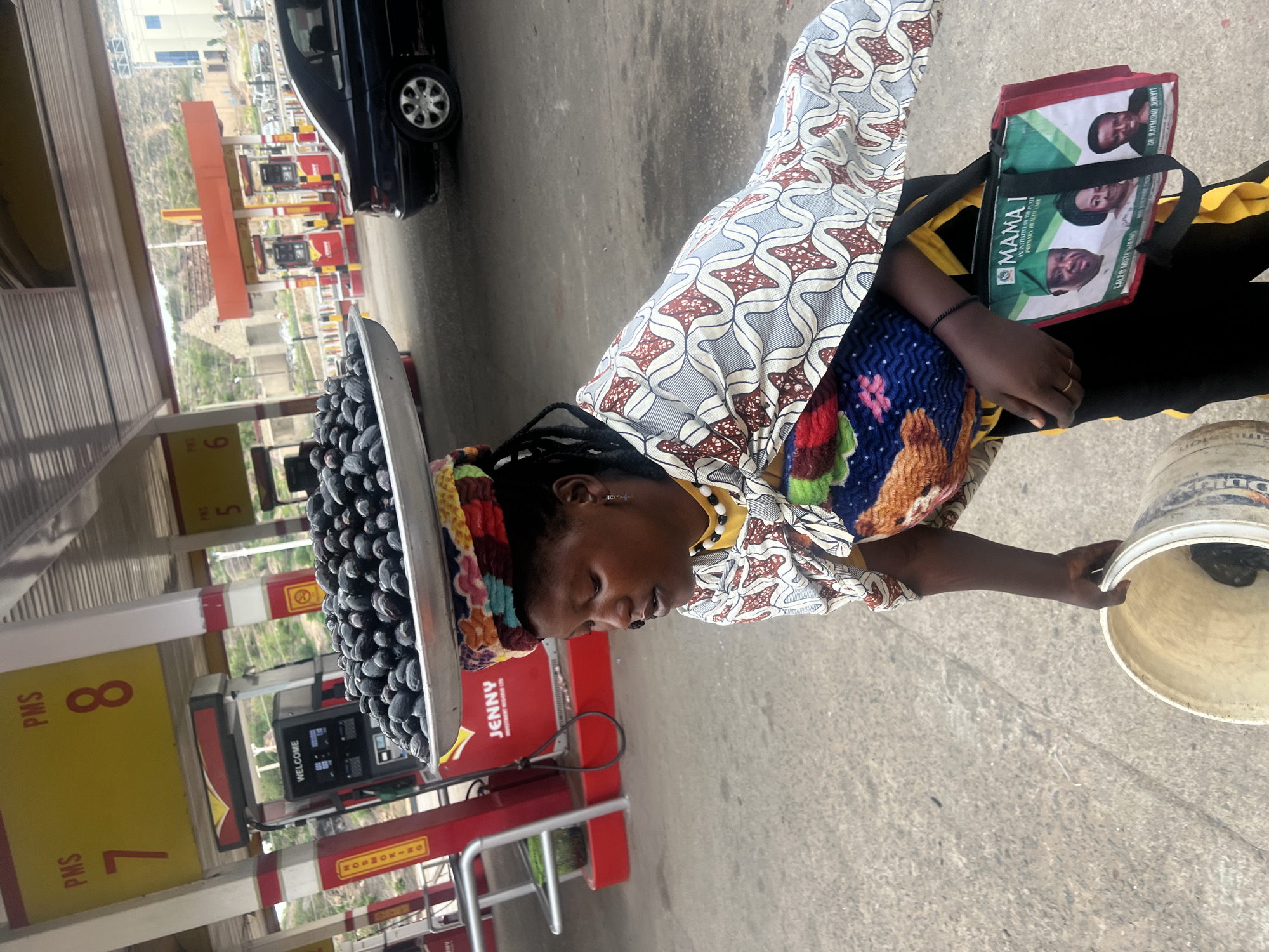 An outdoor scene depicts a woman carrying a silver tray laden with what appear to be dark, round fruits on her head. She is at a gas station in Gura Top, Nigeria. She is facing left, wearing a patterned headwrap, a patterned shirt, and a colorful knitted sash. She has a bag slung over her shoulder that displays images of several men and the text "MAMA I" and mentions "Primary Healthcare." In her right hand, she holds a bucket. Behind her are gas pumps, labeled "PMS 7" and "PMS 8", as well as other pumps labeled 5 and 6. A car is visible in the background. The ground is paved and appears to be concrete or asphalt.