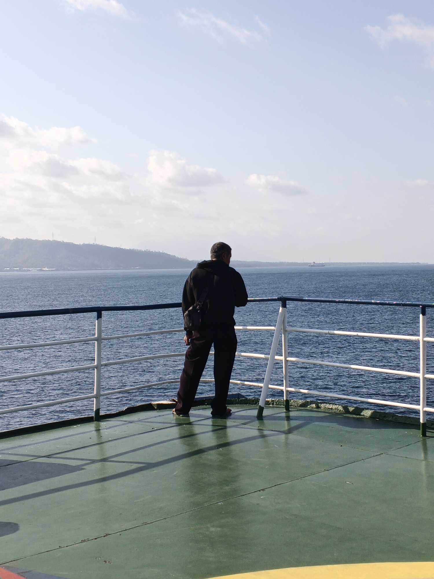 A full-shot photograph captures a lone man standing on the deck of a ferry, looking out at the ocean. The man, facing away from the camera, is dressed in a black hooded sweatshirt and dark pants. He has a dark-colored bag slung over his shoulder. The ferry deck is painted a greenish hue with shadows of the railing cast upon it. Beyond the deck, a blue ocean stretches out to the horizon, meeting a hazy, mountainous coastline. The sky above is a pale blue with scattered white clouds. The overall scene suggests a tranquil journey, possibly in the waters near a coastal city in Indonesia.