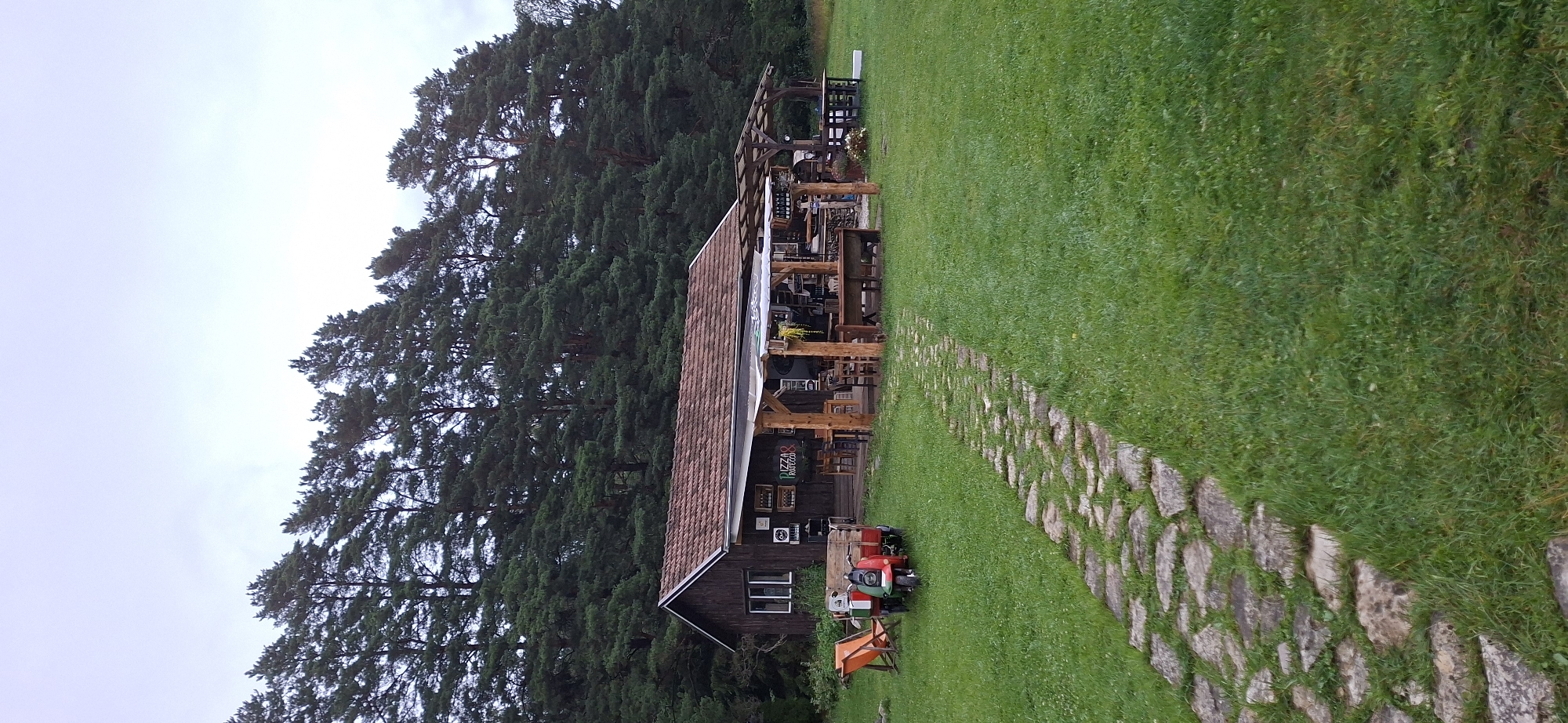 In Orissaare, Estonia, a rustic wooden building, possibly a restaurant, is situated in a grassy field. The building has a tiled roof and a covered outdoor seating area furnished with wooden tables and chairs. In front of the building, a stone pathway leads through the lawn. To the left of the pathway, an orange lounge chair is placed next to a vintage red and green three-wheeled vehicle with a wooden cargo box. A dense forest of tall pine trees forms the backdrop of the scene, and the sky is overcast.