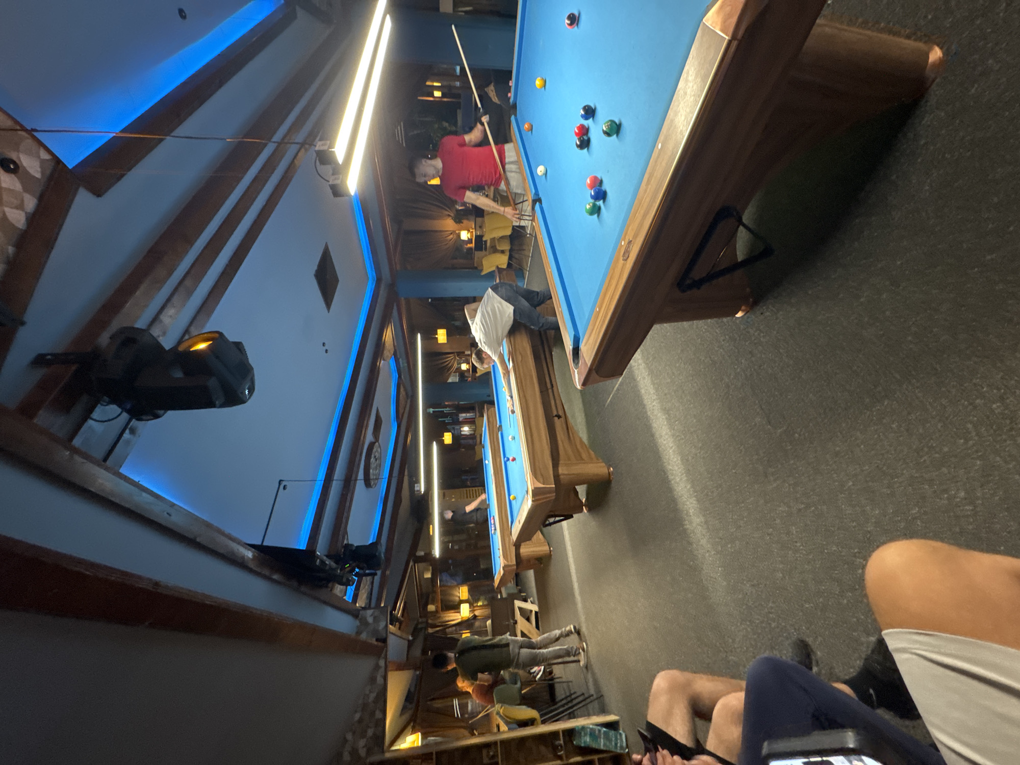 The image depicts the interior of a recreational space with multiple pool tables. Two people are playing pool, one wearing a red shirt and the other a white shirt. Another person is walking in the background. Foreground shows other individuals seated at tables, possibly patrons of the establishment. The space appears to be a pool hall or a bar with pool tables. The ceiling has a modern design with exposed wooden beams and blue lighting. The specific location in Plovdiv, Bulgaria, cannot be determined from this image alone.