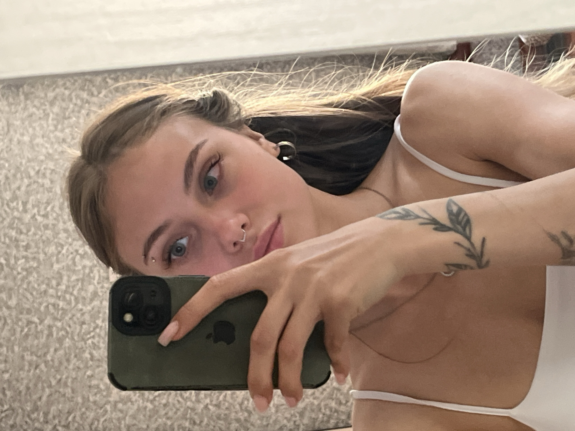 A young woman is captured in a mirror selfie, looking directly towards the viewer with a neutral expression. Her light-colored hair, possibly with blonde highlights, is styled to frame her face. She has prominent blue or light-colored eyes, and her eyelashes are visible. Notable facial piercings include a small stud above her left eyebrow and a V-shaped ring in her nose. A dark, possibly hoop-style earring, is visible in her right ear.
She is wearing a white top with thin spaghetti straps. On her left arm, a tattoo depicting a leafy branch design is visible. In her right hand, she holds a dark green or olive-colored smartphone, which appears to be an iPhone given the visible Apple logo on its back and the camera module design. Her fingernails are painted with a light-colored polish.
The setting appears to be indoors, indicated by the mirror reflection. The background reflecting in the mirror shows a light-colored, textured surface, likely a carpet or rug. Above her head in the reflection, a plain, light-colored wall or ceiling is visible. The lighting is bright and even, suggesting either natural light from a window or well-distributed artificial light. No specific outdoor or architectural cues related to the location context of Фоки, Russia, are visible within this indoor setting.