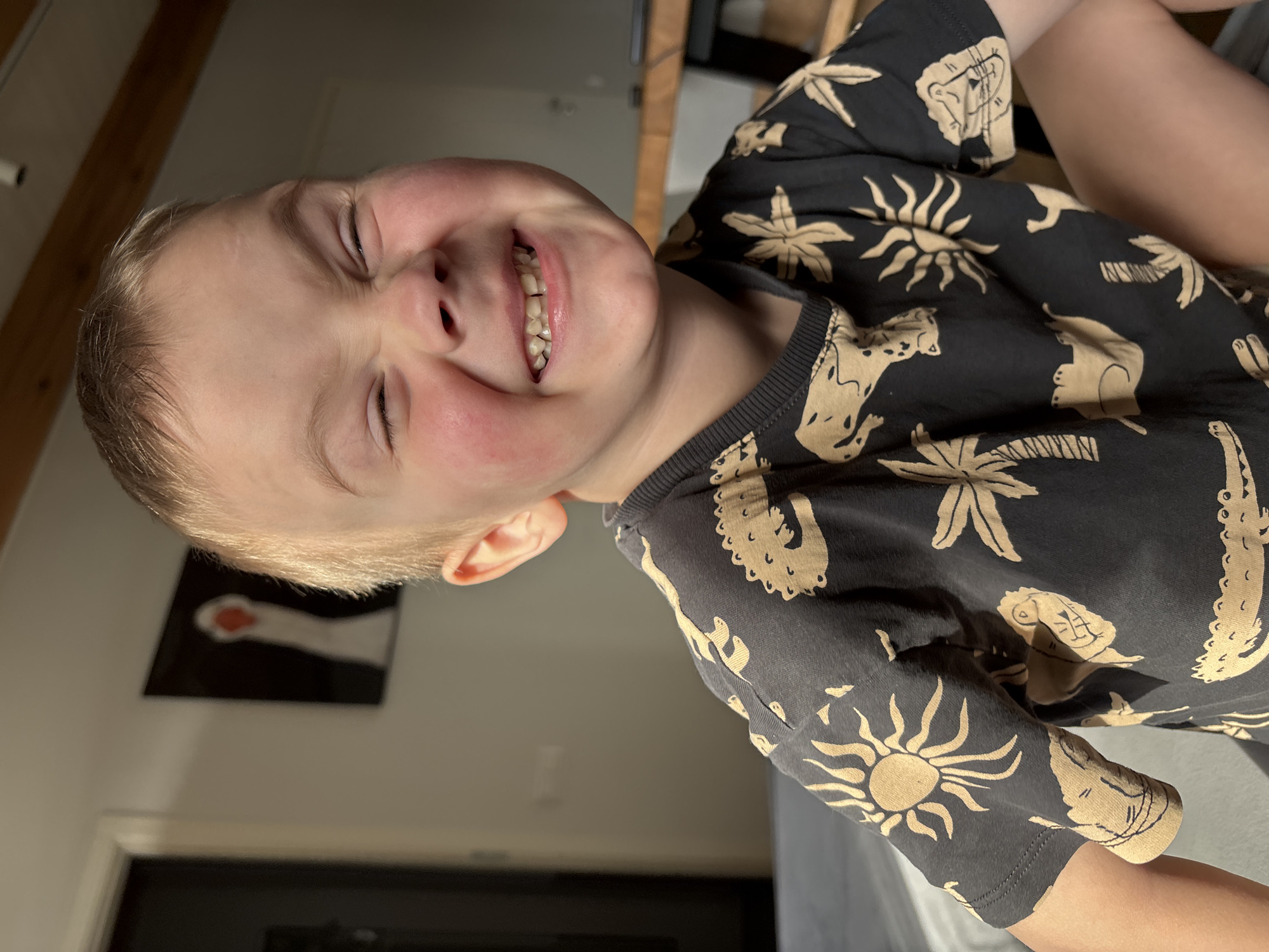 A young boy with blond hair and a black shirt with tan-colored animal and sun designs is smiling broadly with his eyes closed. The background shows a room with a picture on the wall, and natural light is coming through a window. The location is Chaikovsky, Russia.