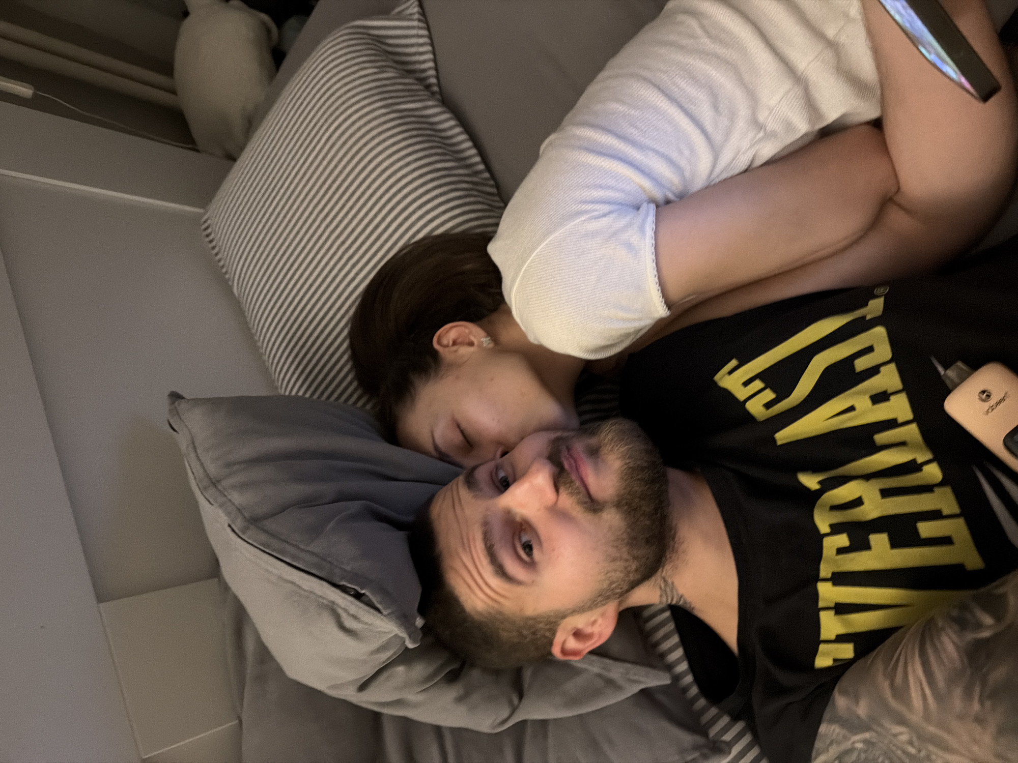 Two people are lying on a bed, presumably indoors. A woman with dark hair is lying on top of a man, with her head near his face, possibly kissing him. The man has short hair and a beard and is wearing a black t-shirt with yellow lettering. The woman is wearing a white shirt. There is a striped pillow behind the woman and a grey pillow under the man's head. A white object is visible in the upper left corner of the image. A phone is visible near the woman's legs. A device of unknown function is on the man's right. The location is unknown and not indicated in the image. No location in Sofia, Bulgaria is evident.