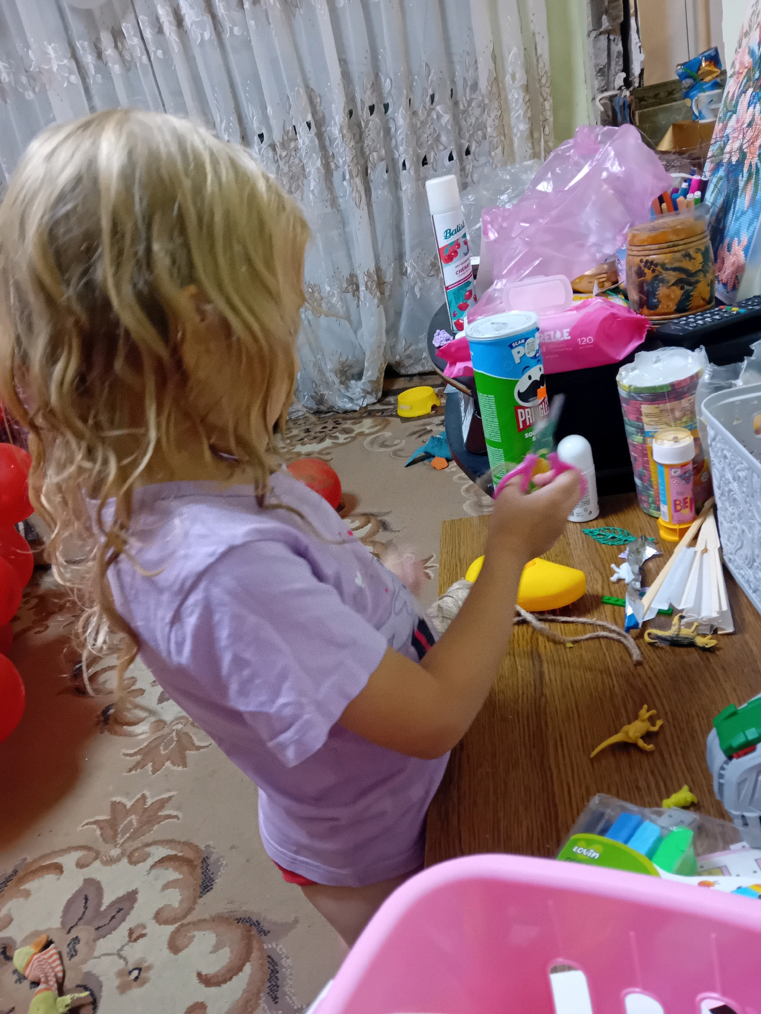 The image shows a young Caucasian girl with blonde hair, seen from behind and from the side, wearing a light purple t-shirt and red shorts. She is sitting at a wooden table, engaged in an activity with scissors in her right hand. The table is cluttered with various items, including a can of Pringles, a can of Batiste dry shampoo, a pack of wet wipes, several cups with colorful contents, a small white spray bottle, a yellow plastic object, a spool of twine, a green rubber band, a small white umbrella, a toy lizard, and a small figurine. In the background, a light-colored curtain with a floral pattern is visible, and red balloons are scattered on the floor. The scene appears to be indoors, likely a domestic setting. There is no discernible information to pinpoint the exact location within Chuhuiv, Ukraine.
