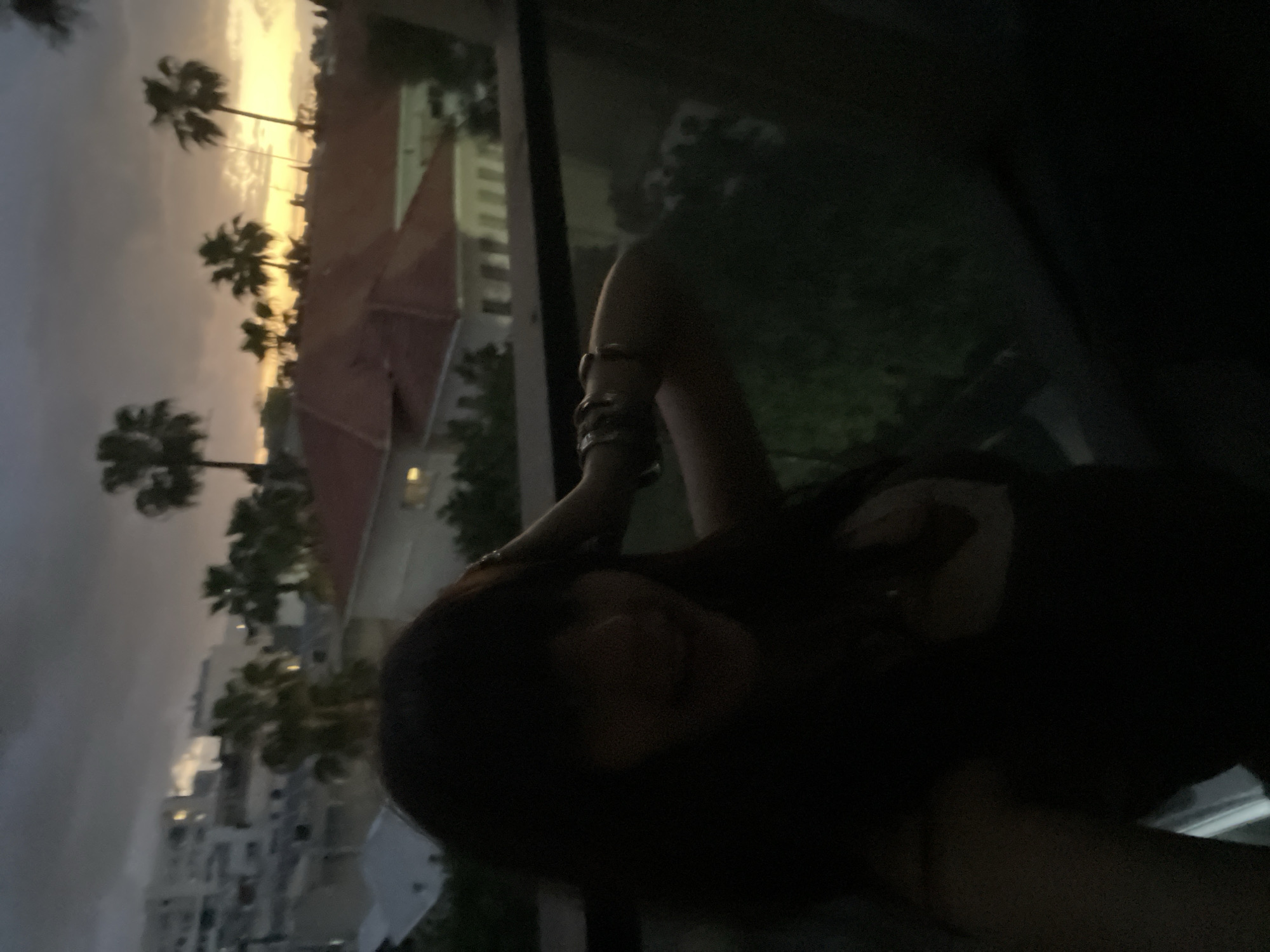 A woman with dark hair is partially visible in the foreground, likely sitting by a window. Her arm is raised, adorned with several bracelets. Outside the window, a cityscape unfolds during the dusk. Tall buildings and palm trees are visible against an orange sky. The scene takes place in Raanana, Israel. The presence of residential buildings and palm trees suggests a coastal or urban environment within the city.