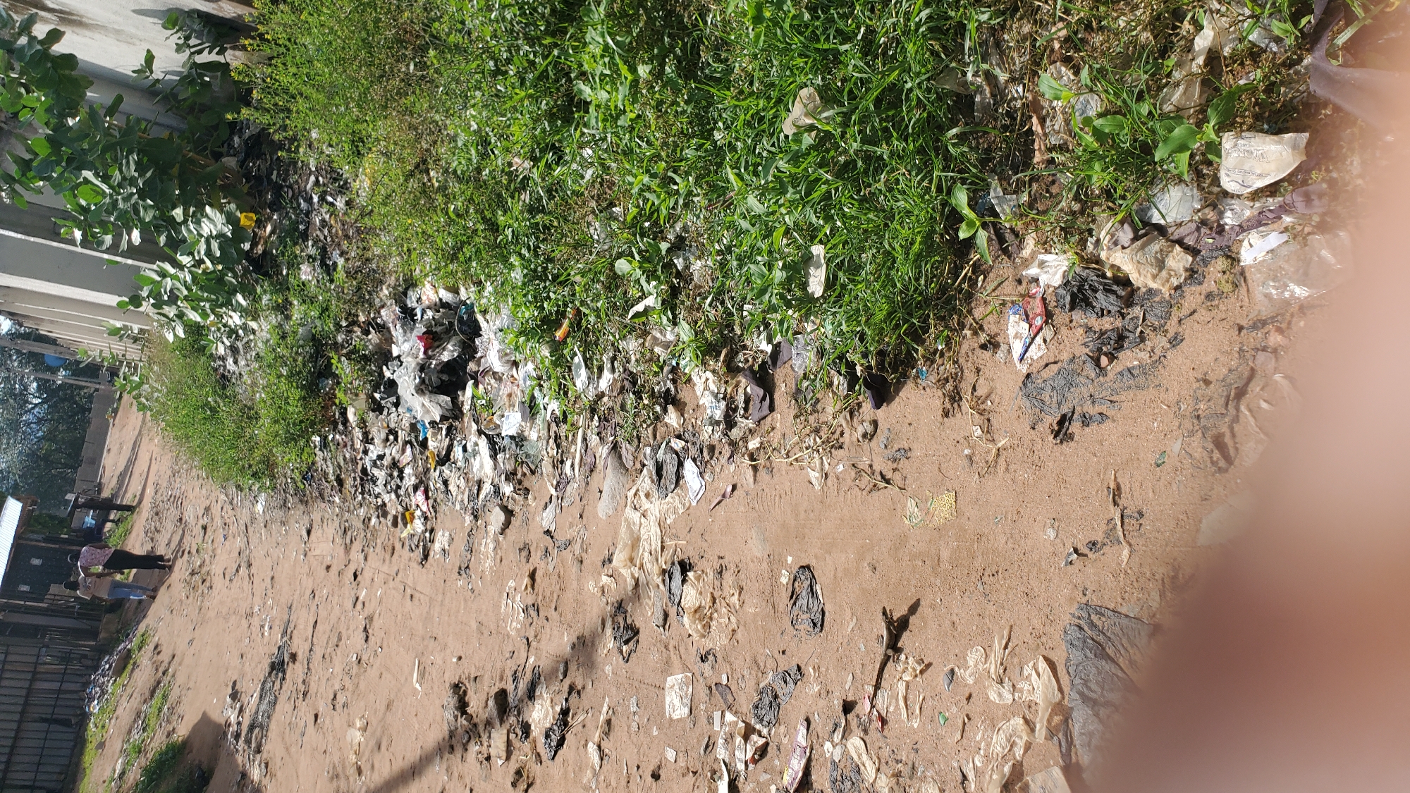 The image depicts a dirt road in Jalingo, Nigeria, heavily littered with trash, primarily plastic bags, wrappers, and other debris. The road is sandy and uneven, with some patches of sparse vegetation growing along the edges. In the background, there are buildings with corrugated metal roofs and a concrete wall. Two individuals, seemingly adult males, are walking away from the camera down the road. One is wearing a light-colored shirt and dark pants, while the other is in a darker shirt and lighter pants. The overall scene suggests an unkempt urban environment with significant waste management issues.