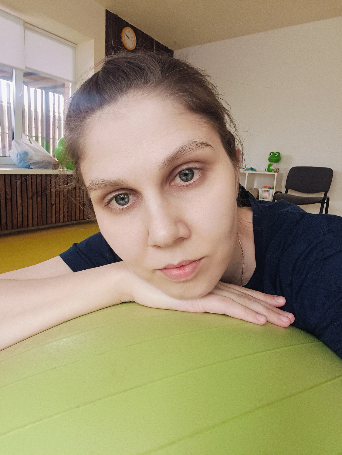 A woman is positioned prominently in the foreground, resting her head and forearms on a large, ribbed, lime-green exercise ball. She has light skin, dark hair pulled back, and light-colored eyes (greyish-blue) with visible dark circles beneath them. She is looking directly at the camera with a neutral, perhaps slightly pensive, expression. She wears a dark blue or black t-shirt and a thin gold chain.
The setting appears to be indoors, likely a room for exercise or therapy. In the background to the left, a large window is visible, letting in bright daylight. Outside the window, a wooden slatted structure, possibly a fence or part of a building, can be seen. A white plastic bag and another green object are on the windowsill. To the upper left, a portion of a yellow wall or floor is visible.
To the right of the woman, the room continues with a plain white wall. On a small white shelving unit against this wall, a green frog figurine is visible. A dark-colored chair with a black frame and seat is positioned further back. Higher on the wall, a round clock with a yellow frame and white face is mounted. The overall lighting suggests it is daytime. There is no visible text in the image.