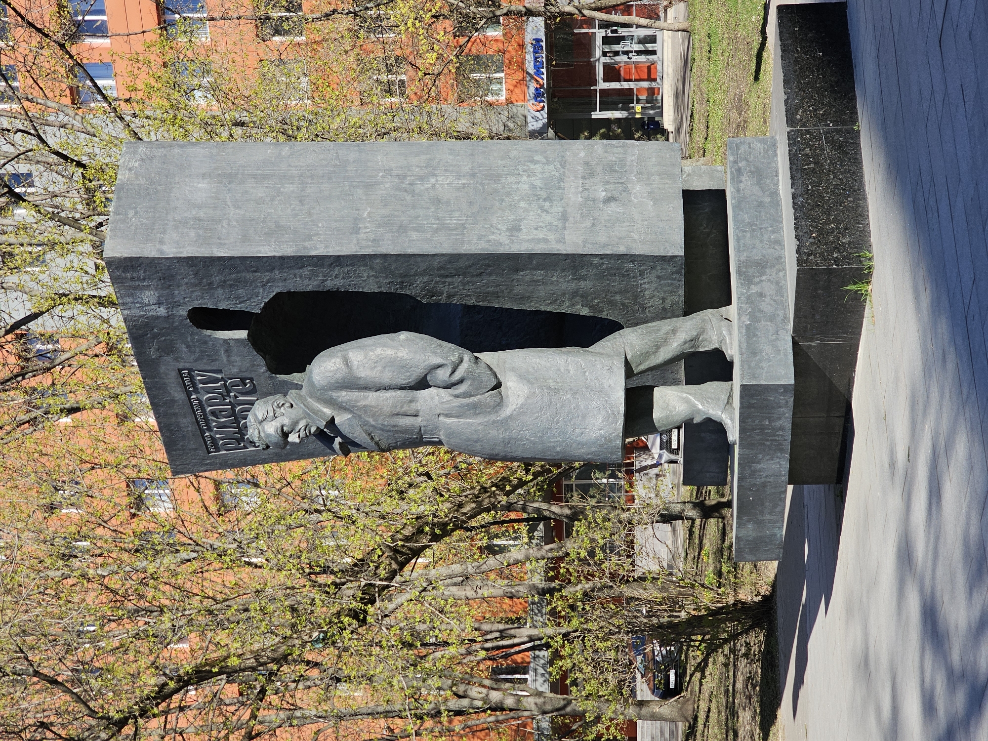 The image shows a statue of Andrei Sakharov in Moscow, Russia. The statue is made of dark metal and depicts Sakharov in a long coat. The statue is located in a park near a building. The park is surrounded by trees and grass. The sky is blue and there are some clouds.