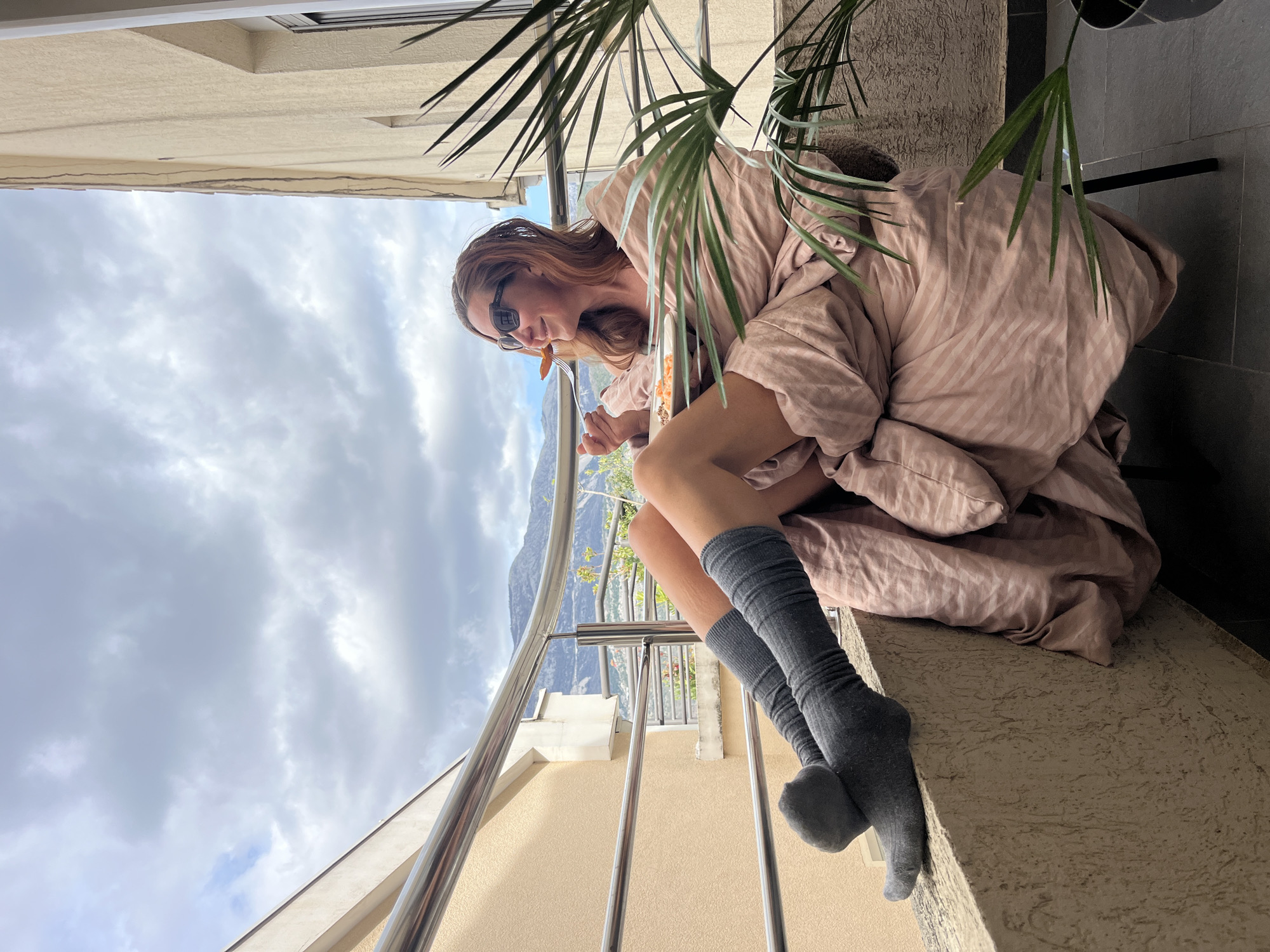 A woman in sunglasses is sitting on a ledge, wrapped in a striped pink blanket, wearing grey knee-high socks. She is eating something from a fork. A railing is in front of her, with a cityscape visible in the background, including mountains and a cloudy sky. Palm fronds are partially obscuring the view. The location appears to be a balcony in Budva, Montenegro.