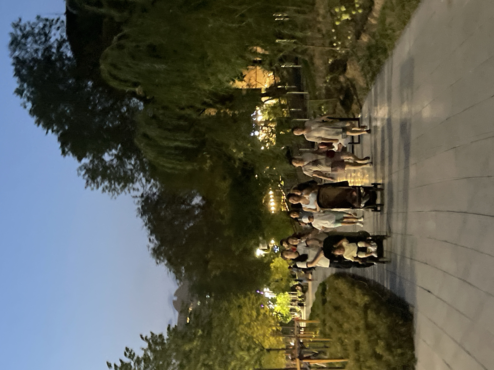 A group of people, including adults and children, are walking on a paved pathway in a park at dusk. The path is lined with low green bushes on the left and trees on the right. In the foreground, a child is seated in a stroller being pushed by an adult. Further down the path, more people are walking, some with strollers. Lights are visible in the background, suggesting urban surroundings. The sky is a pale blue, indicating the transition from day to night. Based on the context provided, this scene is located in the city of Almaty, Kazakhstan.