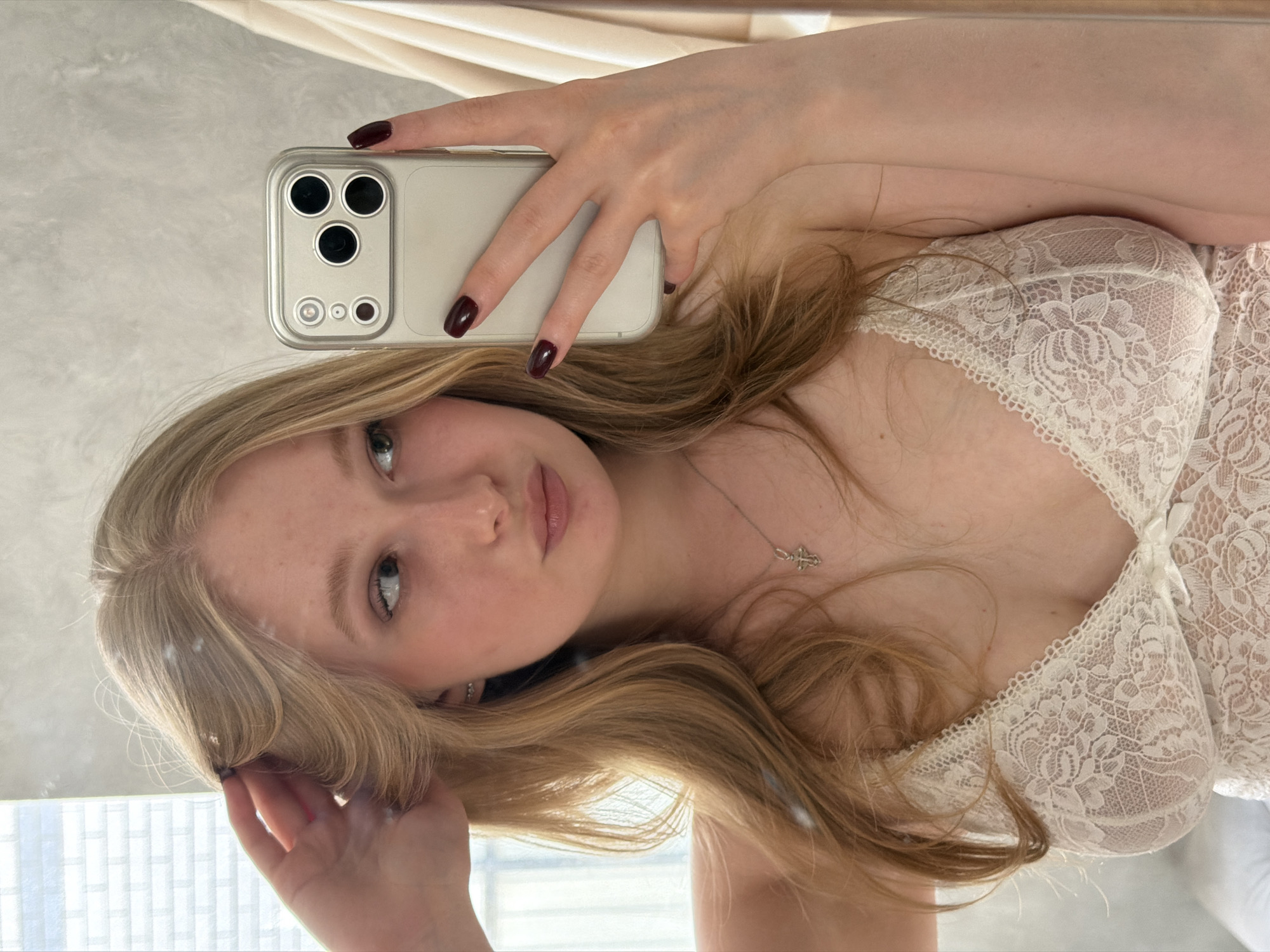 A blonde woman is taking a selfie with her smartphone in Rossiyskiy, Russia. She is wearing a white lace top. Her nails are painted a dark red color. She has a silver necklace with a cross pendant. The background appears to be a room with a window.