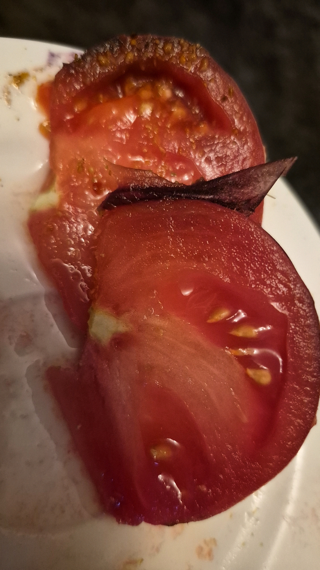 The image displays two slices of a ripe tomato on a white plate. The tomato slices are a vibrant red, with visible seeds and juicy pulp. One slice is positioned above the other, with a piece of dark red leaf-like vegetable or herb resting between them. The background is dark and out of focus, suggesting an indoor setting, possibly a kitchen or dining area. There is no indication of the city of Krauja, Latvia, in the image itself. The scene is a close-up, detailed view of the tomatoes, emphasizing their texture and freshness.