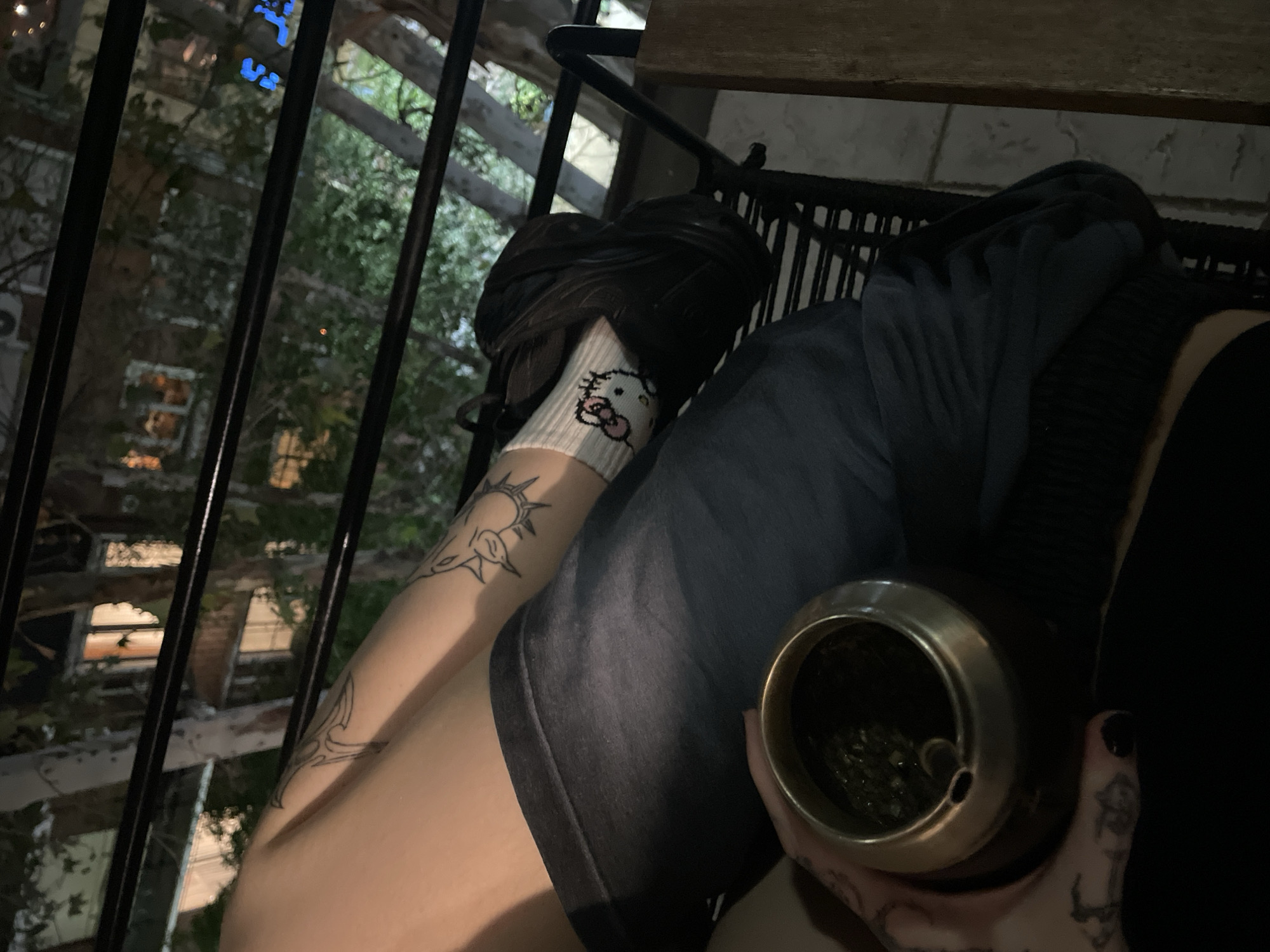 The image shows a person seated, presumably on a balcony or elevated outdoor area, evident by the wrought iron railing on the left side of the frame. Outside the railing, there are multi-story buildings. The person in the image is wearing a dark-colored hoodie or top and denim shorts. Their legs are visible, with a black shoe on one foot and a white sock with a Hello Kitty design. Both legs have tattoos. The person is holding a mate gourd. The environment appears to be urban, possibly in Buenos Aires, Argentina.