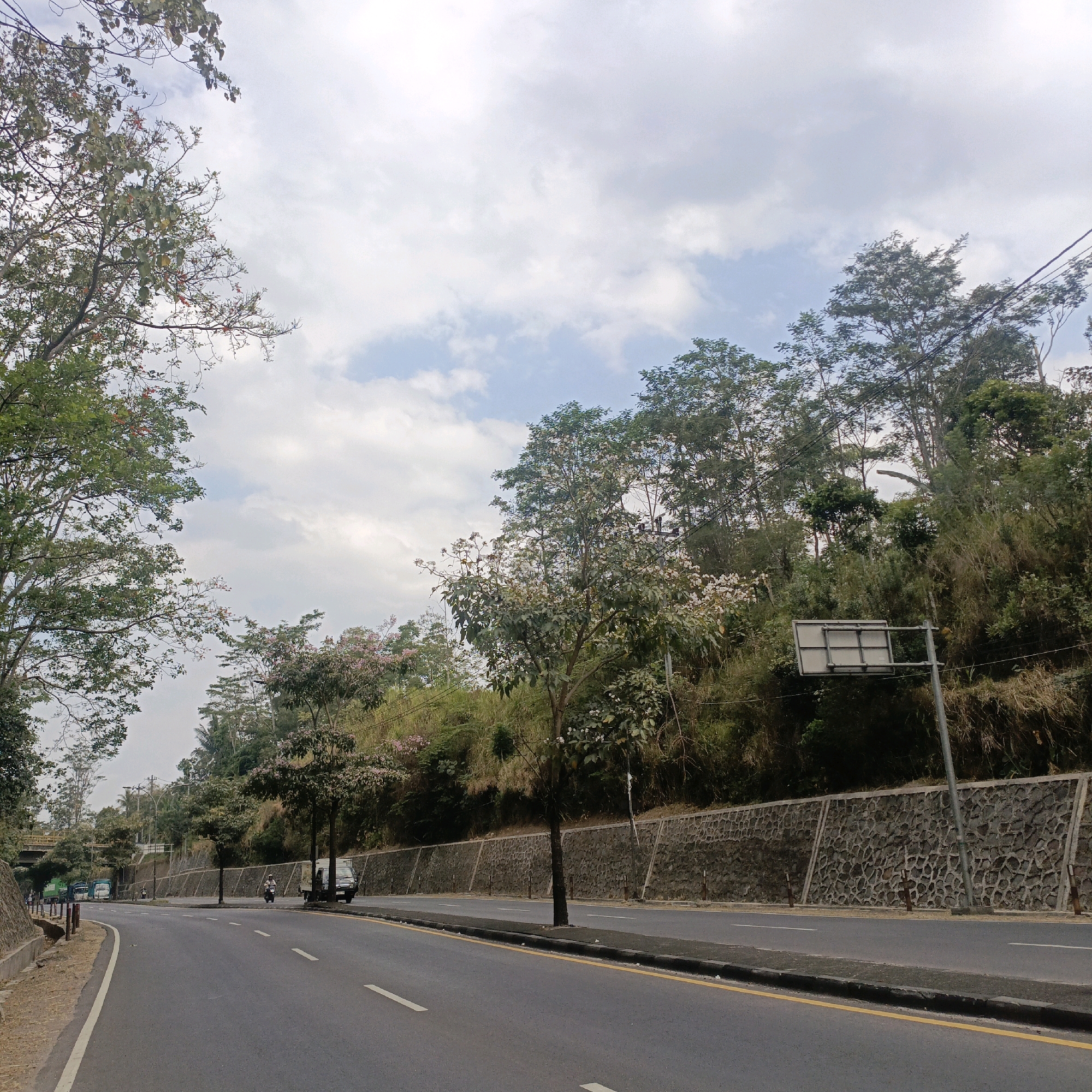 The image depicts a paved road curving through a landscape with trees and a stone retaining wall. The road has white lane markings and a yellow line along the curb. A few vehicles, including a motorcycle, a van, and a truck, are visible on the road. Trees line the roadside, with a dense green hillside beyond the retaining wall. The sky is partly cloudy. The location is identified as a road in Salatiga, Indonesia.