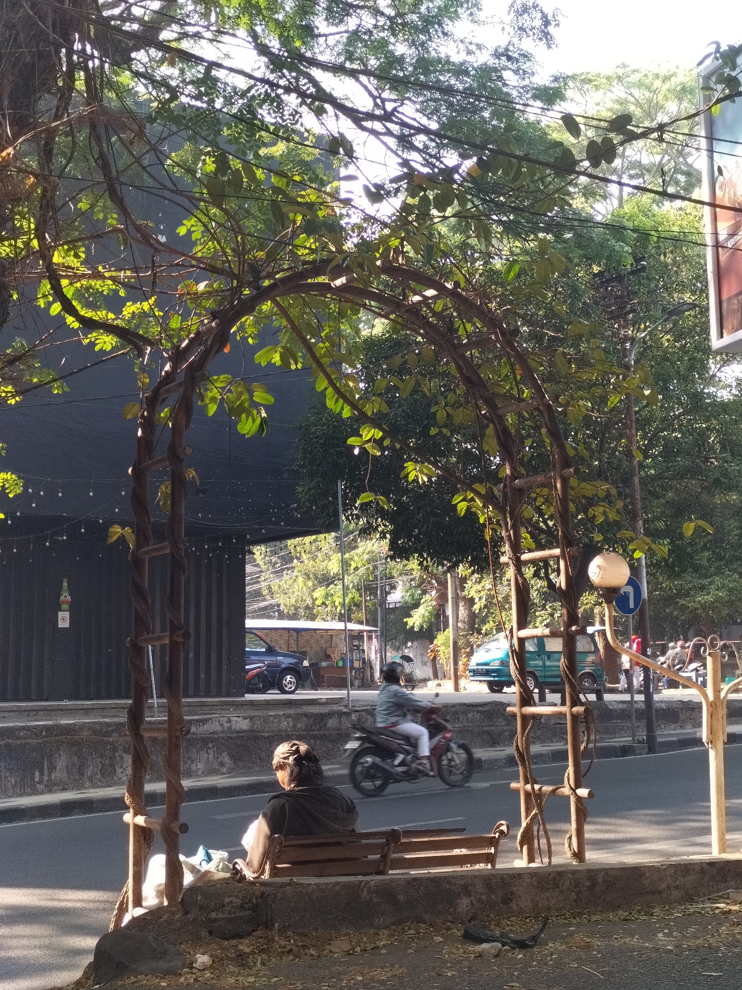 The image depicts a street scene in the city of Malang, Indonesia, bathed in bright sunlight filtering through lush green foliage and tree branches. In the foreground, an arched trellis, seemingly constructed from twisted vines and rustic poles, dominates the view. A person is seated on a wooden bench beneath the trellis, their back to the camera. The bench is positioned on a raised concrete area bordering a paved road.
On the road, a person on a motorcycle is captured in motion, their figure slightly blurred due to the speed. Further down the street, a turquoise van is parked, and a dark blue car is also visible. Behind the road, there's a building with a dark facade, adorned with a string of small lights. The background is filled with dense trees and vegetation, suggesting a tropical environment. A street sign with a blue arrow indicating a turn is present, and a white spherical street lamp on a pole stands to the right of the trellis. The overall impression is a vibrant, sun-drenched urban setting with natural elements integrated into the cityscape.