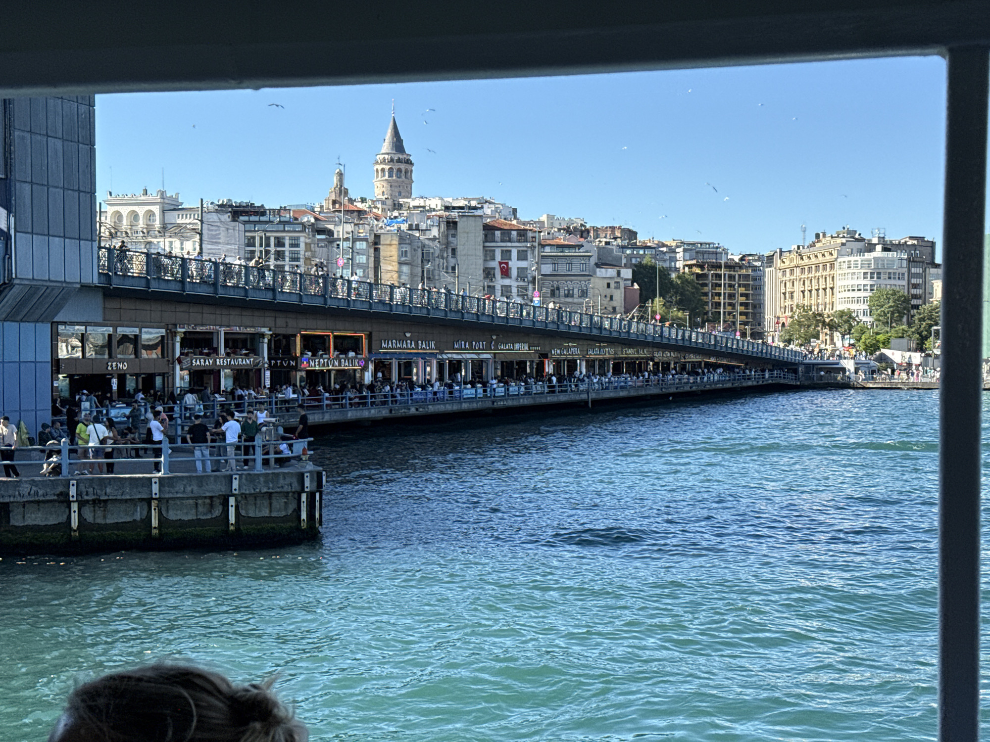 The image shows a view of the Galata Bridge in Istanbul, Turkey, with the Galata Tower visible in the background. The bridge is crowded with people on the upper level, and numerous restaurants line the lower level, including businesses named MARMARA BALIK, MIRA PORT, NEW GALAPERA GALATA, ISTANBUL, ZENO, SARAY RESTAURANT, and NEPTUN BALIK. The body of water between the camera and the bridge is visible and appears to be the Golden Horn or Bosphorus. The picture seems to be taken from inside a boat, with part of the boat's frame visible in the lower right and upper border. There are some buildings, including houses and office buildings, behind the Galata Bridge. Some birds are flying in the blue sky above the buildings. The day is bright and sunny.