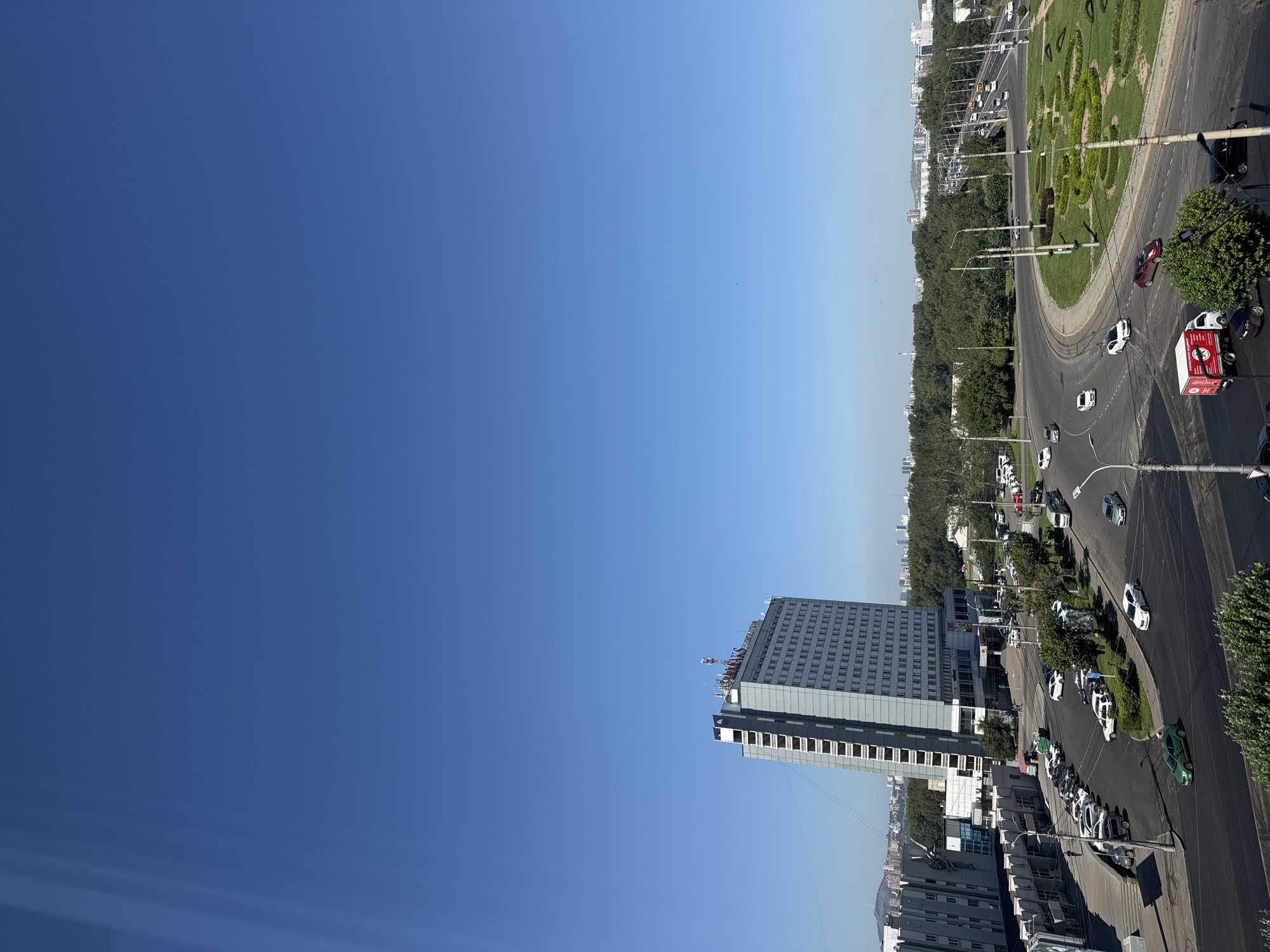 The image depicts a cityscape in Krasnoyarsk, Russia, dominated by a clear blue sky. In the foreground, a modern, multi-story building with a distinctive grey facade and numerous windows stands prominently. The building has signage at its rooftop. Surrounding the building is a network of roads with several cars in motion and parked. A distinct rotary or roundabout with manicured green landscaping is visible, along with streetlights and trees. In the mid-ground, a dense line of trees forms a verdant backdrop. In the distance, other city buildings and infrastructure are visible, suggesting an urban environment. The overall scene captures a daytime view of a functional city area with a mix of residential or commercial buildings and vehicular traffic.