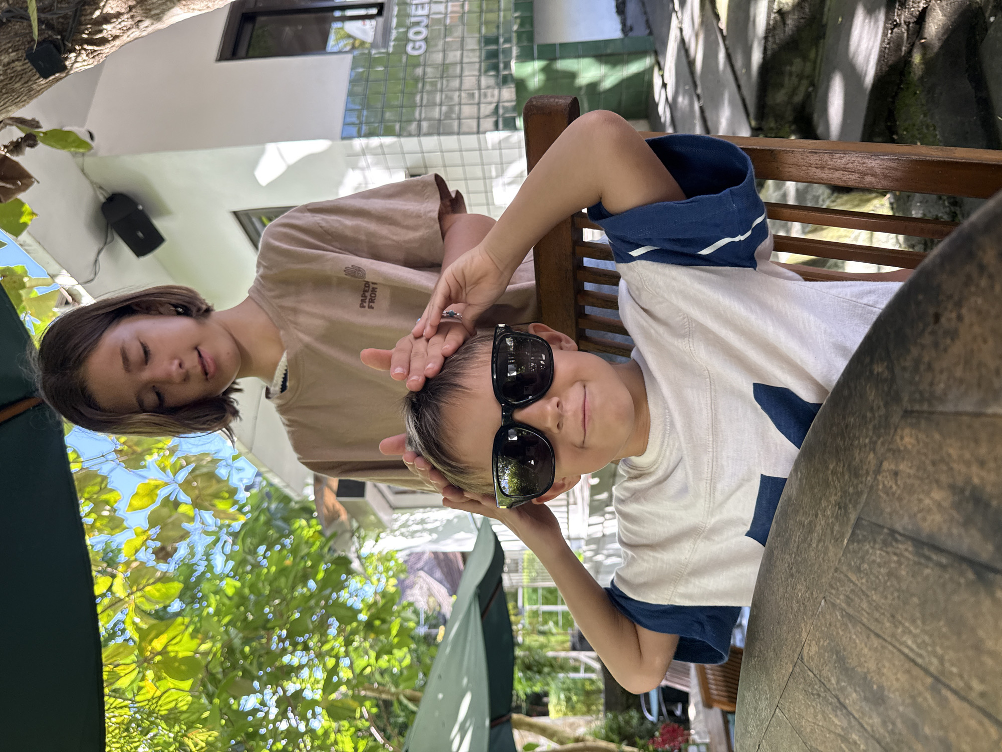In Kuta Selatan, Indonesia, a woman in a tan shirt is touching the head of a child wearing sunglasses while sitting in a wooden chair at a wooden table. The location appears to be outdoors, surrounded by trees and foliage. A building with a sign that reads "GOJEK" is partially visible in the background.