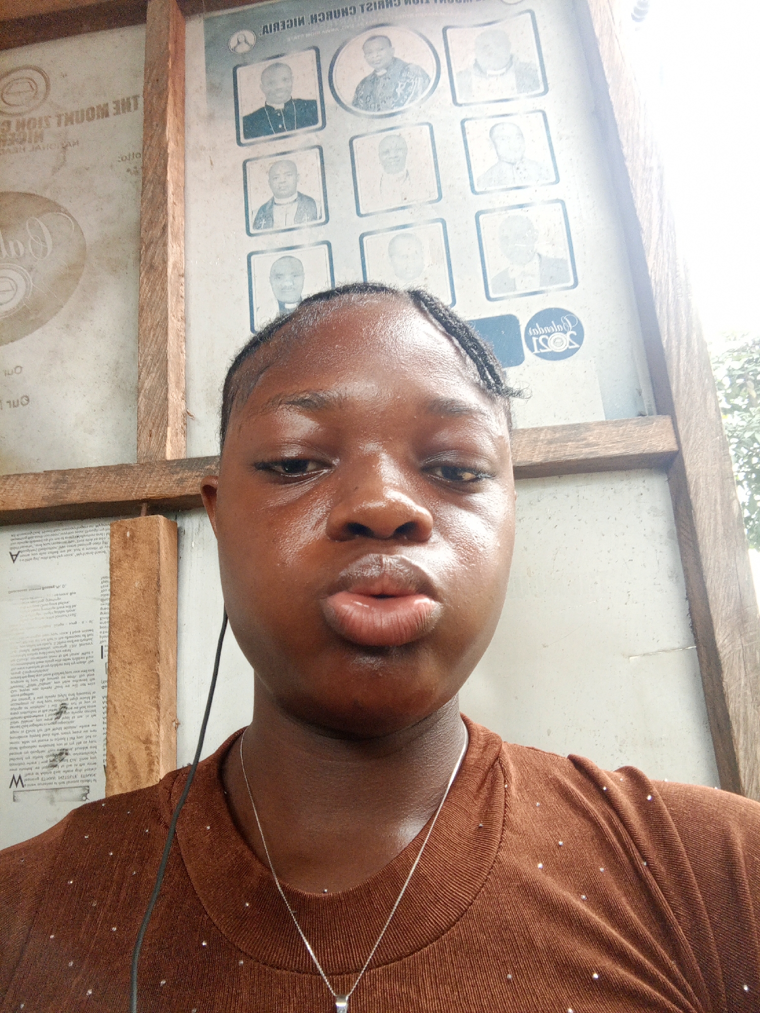 A close-up shot features a dark-skinned woman with braids, a silver necklace, and a brown shirt. Her lips are slightly puckered. In the background, a bulletin board with portraits and text is visible, along with wooden framing. Based on text in the background, it appears that she is standing by the THE MOUNT SION CHURCH NIGERIA, possibly in the city of Mbribit, Nigeria.