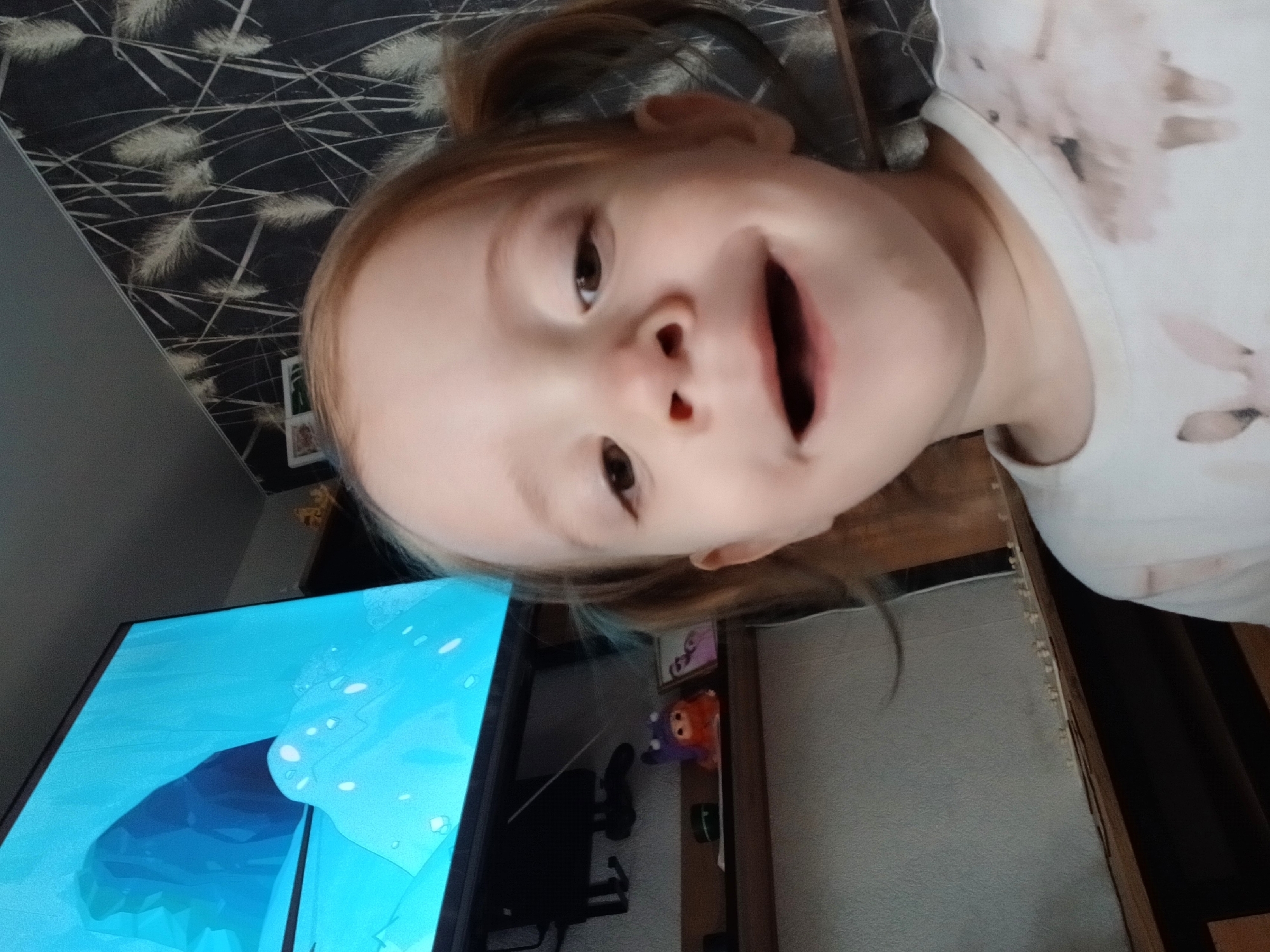 A child with red hair is looking at the camera and has her mouth open, in what appears to be a selfie. She is wearing a white shirt with light pink designs on it. The background includes a TV screen showing a cartoon, a patterned wallpaper, a desk, and wall decor. There is no verifiable evidence to suggest that this image was taken in the city of Марковский, Russia.