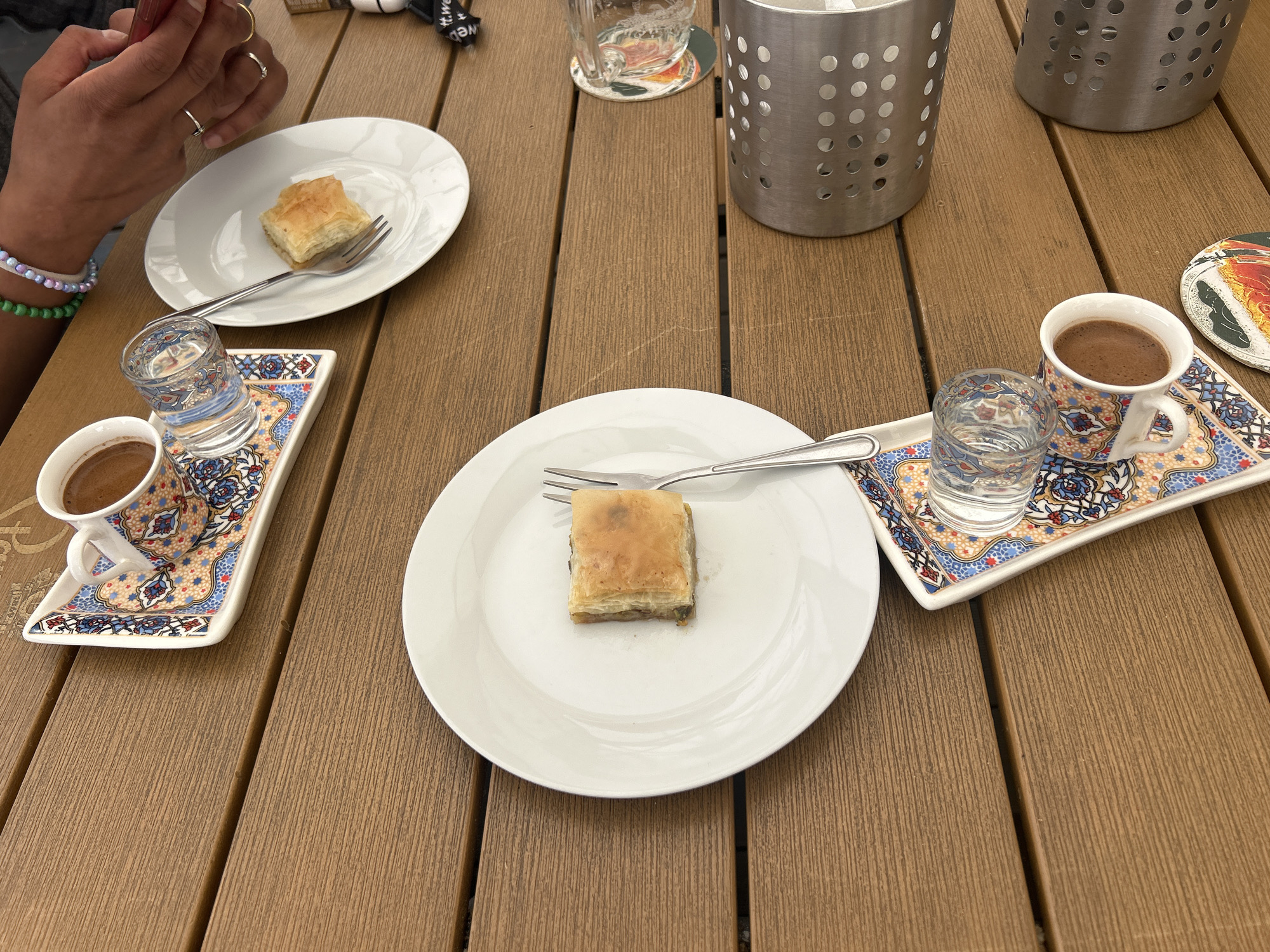 This medium close-up photograph captures an outdoor table setting, likely at a cafe or restaurant in Liberec, Czechia. The scene features three servings of baklava and Turkish coffee on a light brown, slatted wooden table.
In the foreground and center, a square piece of baklava is presented on a white oval plate with a silver fork. To the left, a person's hands are visible, holding a red smartphone. This individual wears several rings and two beaded bracelets (one in white and purple, another in green). Adjacent to their hands is one of three traditional Turkish coffee sets, comprising a small patterned cup filled with dark coffee and a tiny glass of water, all placed on an ornate, rectangular tray with colorful geometric patterns. A similar coffee set and another baklava plate are arranged on the right side of the table.
Two empty, clear drinking glasses are visible, one in the upper middle and another beside the right coffee set. In the background, two cylindrical silver utensil holders with perforated sides stand on the table. A small, circular coaster with an abstract design is partially visible on the far right. A small black item near the top center shows partially obscured white text, reading "t.we" and what might be the beginning of another word like 'b' or 'a'.
The activity depicted is a casual break or social gathering, with individuals enjoying dessert and coffee. One person is engaged with their mobile phone. The lighting suggests daytime, with soft illumination indicative of an overcast sky or shaded outdoor area. The overall ambiance appears relaxed and pleasant, suited for outdoor dining.