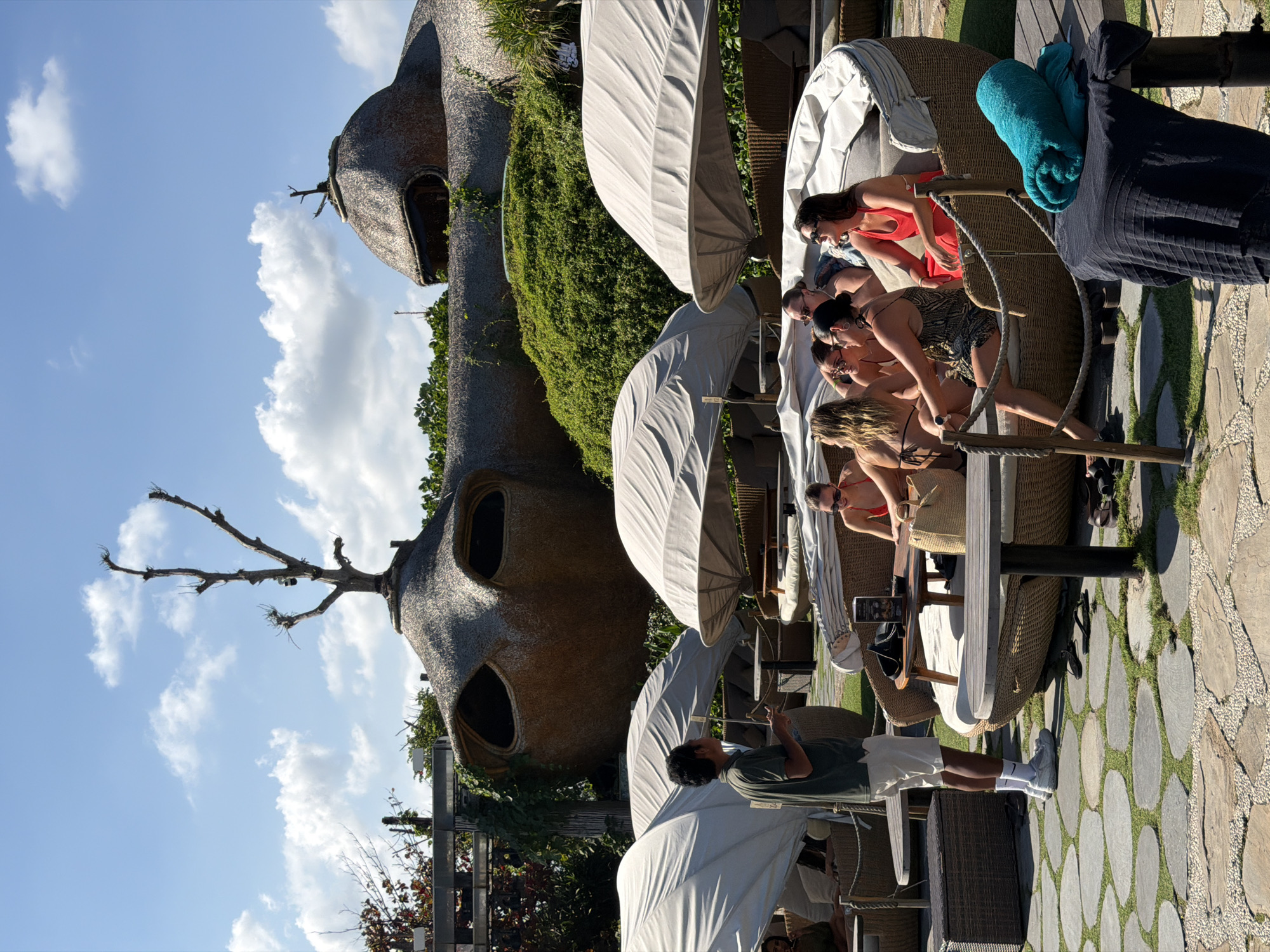 The media file depicts a group of people relaxing in an outdoor seating area near a distinctive, dome-shaped building with large, dark oval openings. The building, covered in a thatched material and overgrown with greenery, appears to be the central architectural feature of the scene. Several individuals are seated in cushioned lounge chairs, some under white canopies, while others are seated at tables.
One woman in the foreground, wearing a red bikini top and sunglasses, is looking to her left. Another woman beside her is wearing a patterned swimsuit. A man in a dark green t-shirt and white shorts stands near a table, looking at his phone. A turquoise towel is draped over the arm of a lounge chair. The ground is paved with irregularly shaped stones and patches of grass. The scene is illuminated by bright sunlight, suggesting it is daytime, and the sky is partly cloudy with a bright blue hue. The overall atmosphere seems relaxed and leisurely. The location is Kediri, Indonesia.