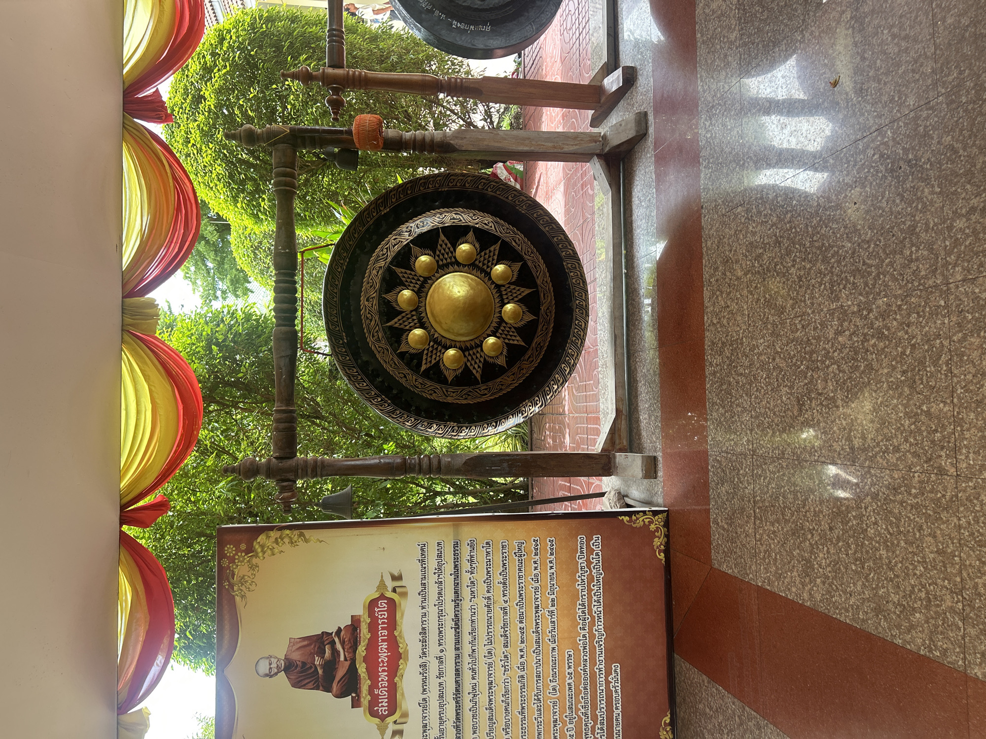 An outdoor shot shows a large, ornate gong suspended from a wooden frame. The gong is dark with intricate gold patterns, and a prominent gold disc at its center. Beside the gong is a framed poster depicting a seated Buddhist monk. The poster has Thai text. In the background, lush green trees and foliage are visible, suggesting a tropical or subtropical environment. Red and yellow drapes are strung across the top of the frame, adding a decorative element. The ground is tiled in shades of brown and beige. The scene is likely located in an outdoor public space in Thai Ko Subdistrict, Thailand, possibly a temple or a cultural site.