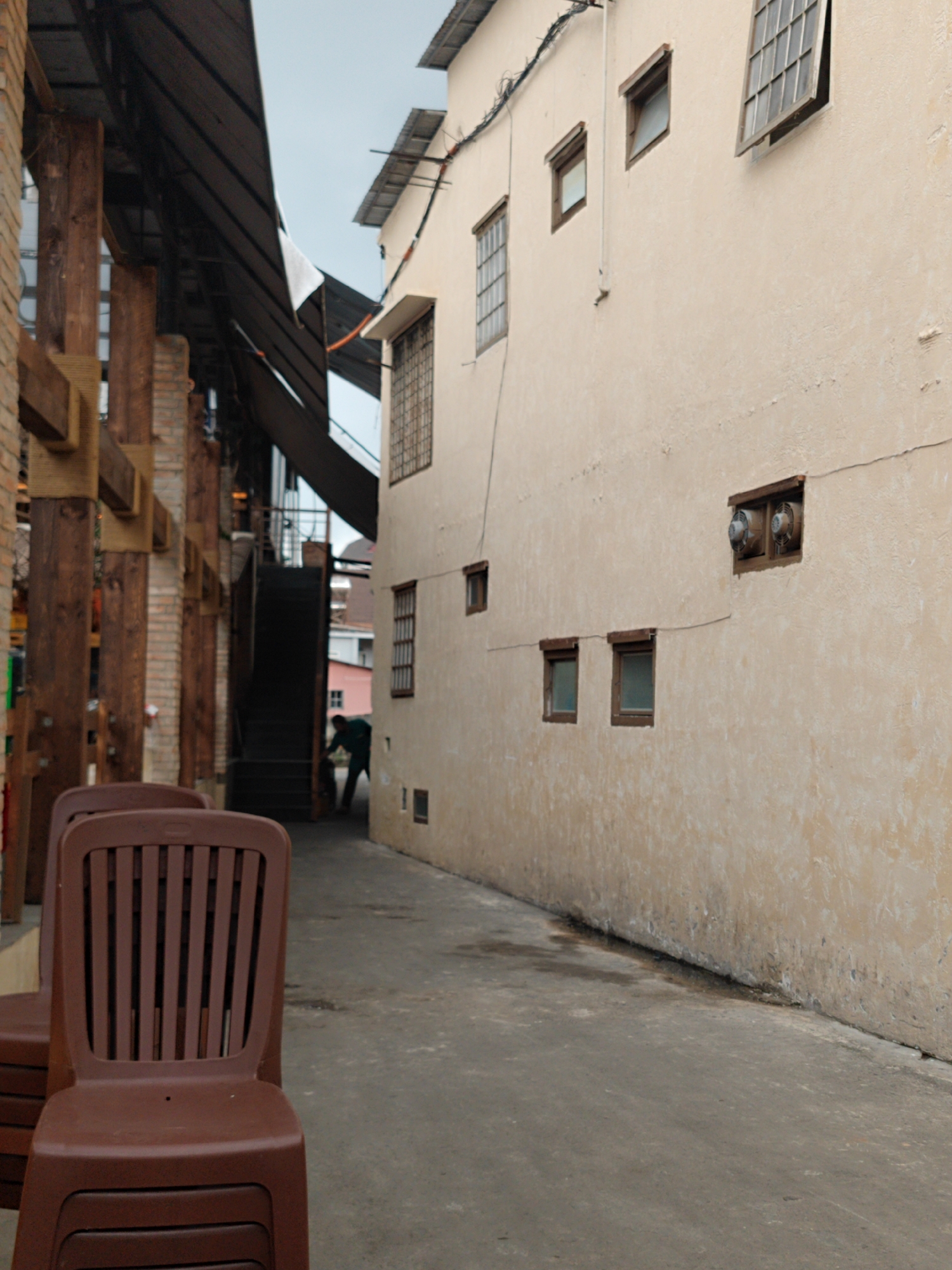 The image depicts a narrow alleyway or passage between two buildings, suggesting a utilitarian or back-of-house area, likely in Cam Ly - Đà Lạt, Vietnam.
In the foreground, on the left side of the concrete path, several brown plastic chairs are stacked. The topmost chair is prominently featured, showing its slatted back and brown hue.
To the left, a structure composed of large wooden beams and brickwork stands, supporting a dark, sloping roof, possibly made of corrugated metal. A small white piece of fabric or plastic dangles from this roof. Further back, a dark staircase ascends, leading out of view. A person, clad in a dark green top and light-colored trousers, is partially visible near the base of these stairs, bending over as if engaged in an activity like cleaning or maintenance.
The right side of the image is dominated by the tall, light cream-colored wall of another building. This wall features several windows of different sizes and designs: some are small and rectangular, one has a protective metal grate, and a larger window is open at an upward angle, revealing a grid pattern. Further along the wall, a recessed opening houses two circular vents. Electrical wires are visible running along the upper part of this building. The wall exhibits signs of age and wear, including cracks and some dampness or discoloration near its base, where it meets the ground.
The ground is a flat, grey concrete or asphalt surface, appearing somewhat grimy. The sky is overcast, providing soft, diffused light, indicating daytime without direct sunlight. There is no visible text in the image. The scene conveys a quiet, functional atmosphere.