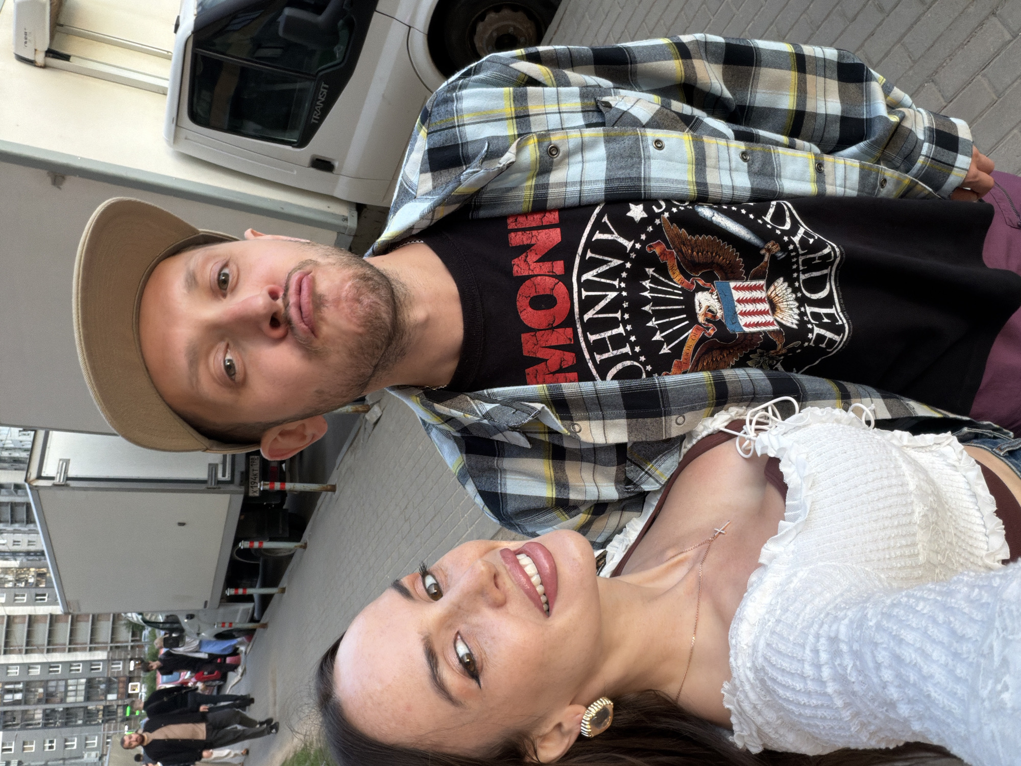This image captures a selfie taken by a couple outdoors, with a blurred urban background suggesting a street scene in Saint Petersburg, Russia. A man, wearing a beige baseball cap and a plaid shirt over a black t-shirt with a graphic of an eagle and the words "MONI JOHNNY", is on the right. He is making a duck face. A woman, on the left, is smiling and wearing a white ruffled top and gold earrings. Behind them, several people are walking along a sidewalk next to parked trucks and buildings. The ground is paved with grey bricks.