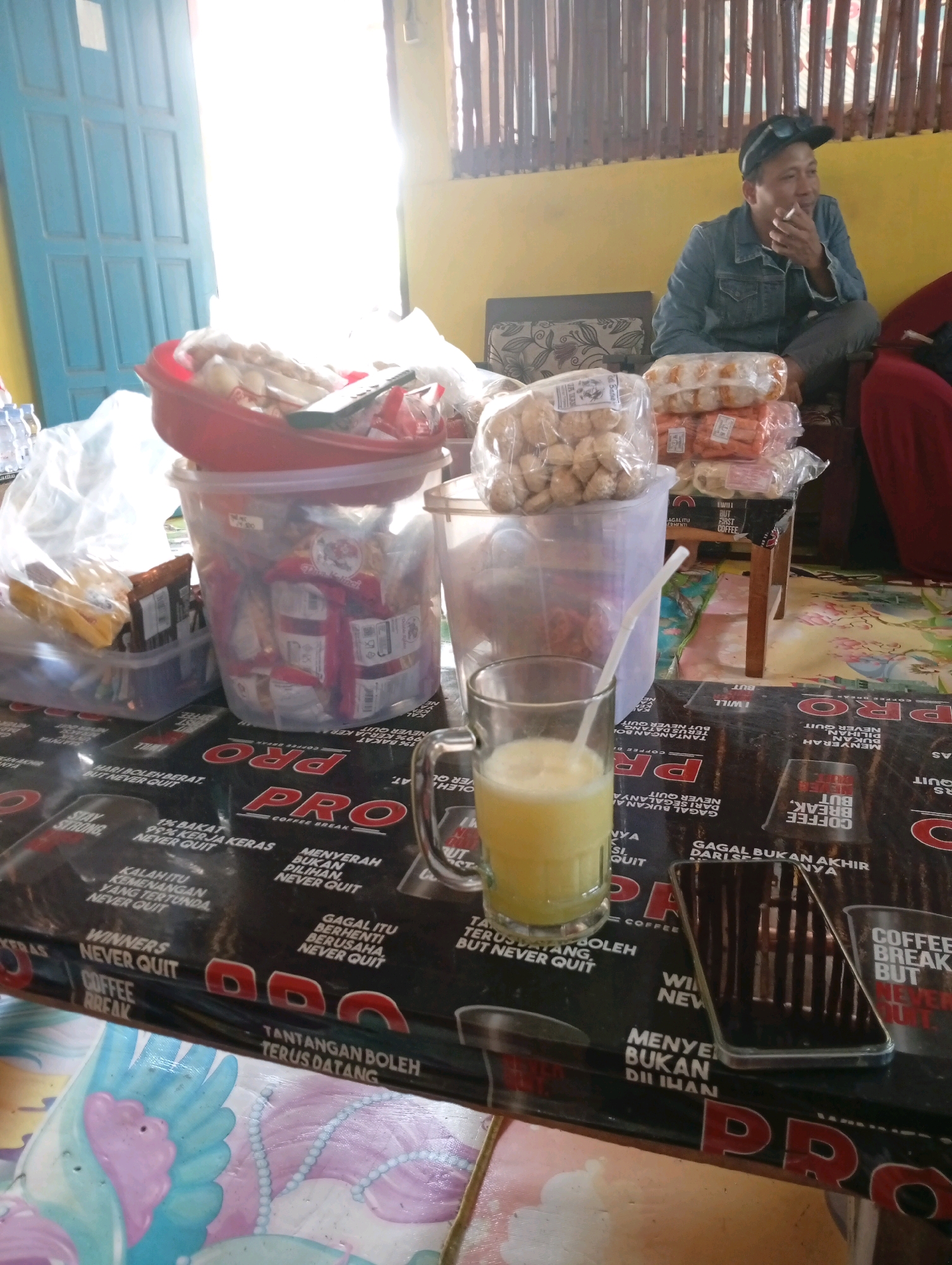 A man wearing a denim jacket and a cap sits with his legs crossed, smoking a cigarette. He is in an indoor setting with yellow walls and bamboo-like structures on the upper wall. In front of him, on a table, are several clear plastic containers filled with food items, possibly snacks or pastries. There is also a glass of yellow liquid with a straw, a smartphone, and various food packaging. The table surface is covered with black material featuring text and logos, including "PRO COFFEE BREAK". The overall impression is a casual gathering or break in a somewhat cluttered but possibly open-air environment. The specific location in Boyolangu, Indonesia, is implied by the context but not visually confirmed in the image itself.