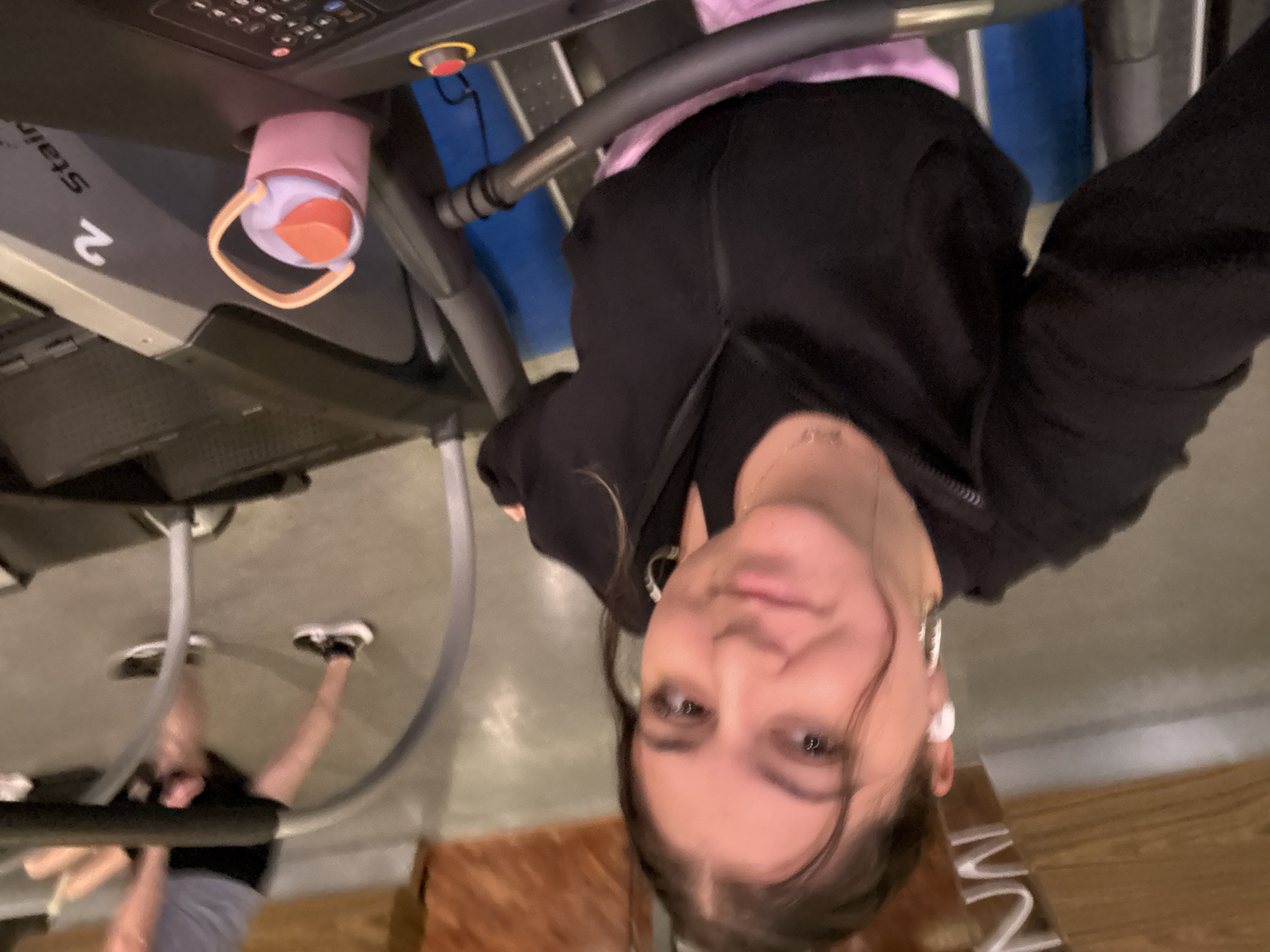 The image, rotated 90 degrees counter-clockwise, depicts a young woman taking a selfie in what appears to be an indoor gym or fitness center. She looks directly at the camera with a neutral to slightly smiling expression. She has dark hair, dark eyes, and is wearing a black zippered hoodie over a light pink or purple top. A white earbud is visible in her right ear.
In the foreground, a treadmill is prominent. Its control panel features various buttons, a digital display, and a red emergency stop button. A pink and orange water bottle with a handle is placed in a cup holder on the treadmill. The side of the treadmill clearly shows the number "2" and partially visible text that appears to read "Stair".
In the background, behind the main subject, another person or a reflection is visible, also seemingly on exercise equipment, identifiable by their legs and white athletic shoes. The floor is a light gray. Sections of a blue wall and a wooden-patterned surface are also visible. Indistinct, partially visible text appears on the wooden-patterned surface in the lower right. While the general setting is a gym, there are no specific visual cues within the image itself to indicate the provided context of Mexico City, Mexico.