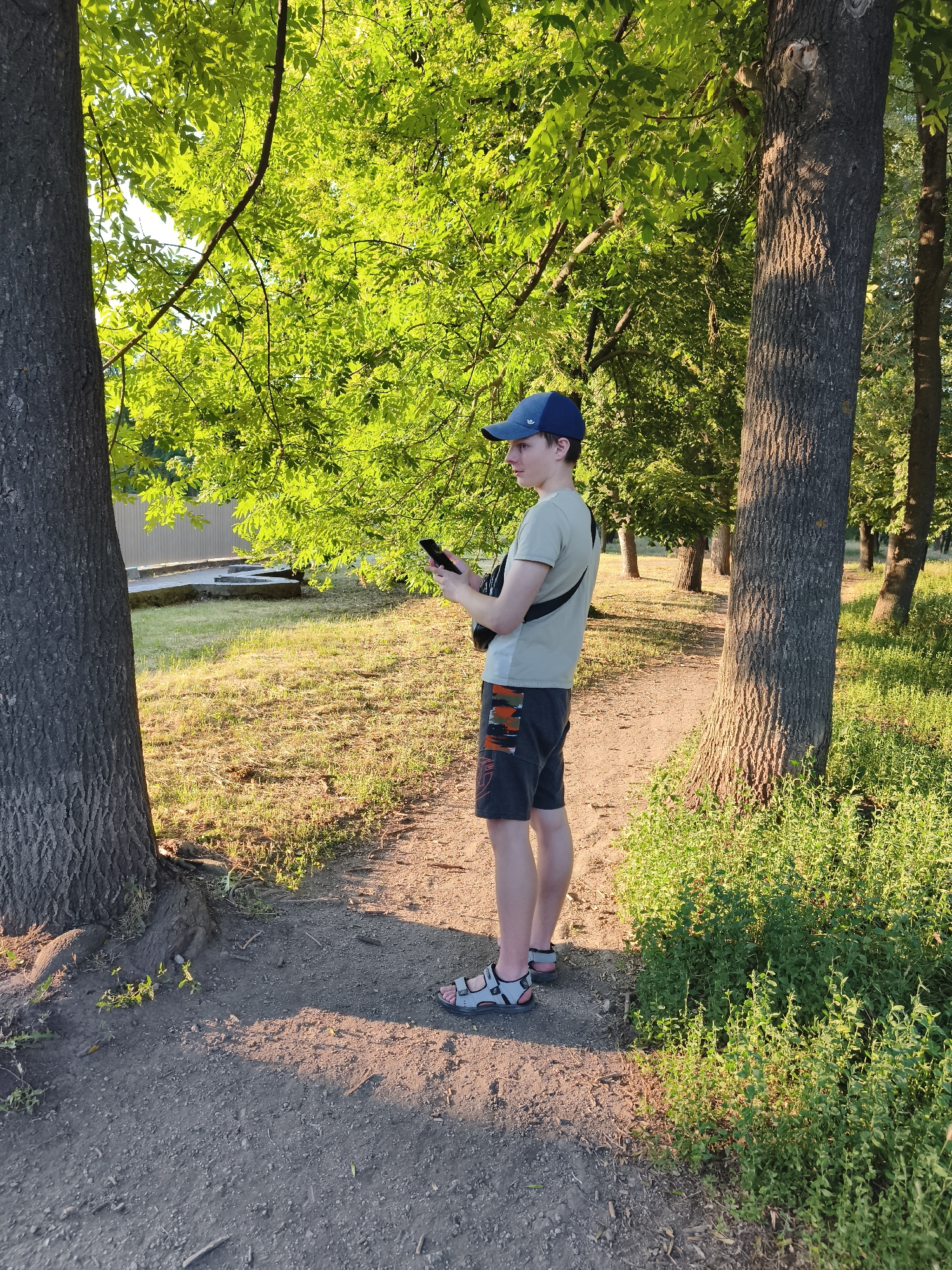 A young male wearing a light-colored t-shirt, dark shorts with a camouflage pattern, a blue baseball cap, and sandals stands on a dirt path in a park. He is holding a smartphone in his hands, looking down at it. A black strap bag is slung across his chest. The scene is set in a park with large trees, lush green foliage, and grass. The sun is casting long shadows, indicating it is late afternoon or early evening. The weather appears to be warm and sunny. In the background, a light-colored fence and some concrete structures are visible. The overall mood is relaxed and casual. The word "UNMATCHABLE" and possibly "LIMITS" are visible on his shorts.