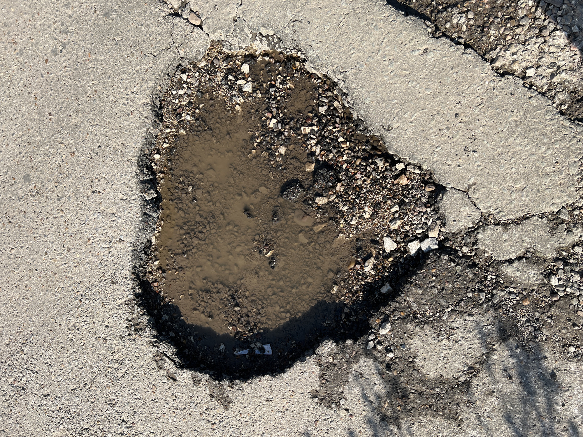 The image is a close-up, overhead view of a heart-shaped pothole filled with muddy water on a cracked asphalt road. The pothole is surrounded by broken asphalt and gravel. The lighting suggests it is daytime, with shadows visible on the road. There are no people or vehicles present in the image. The setting is an urban environment, likely a street in Salavat, Russia, characterized by its damaged road surface.