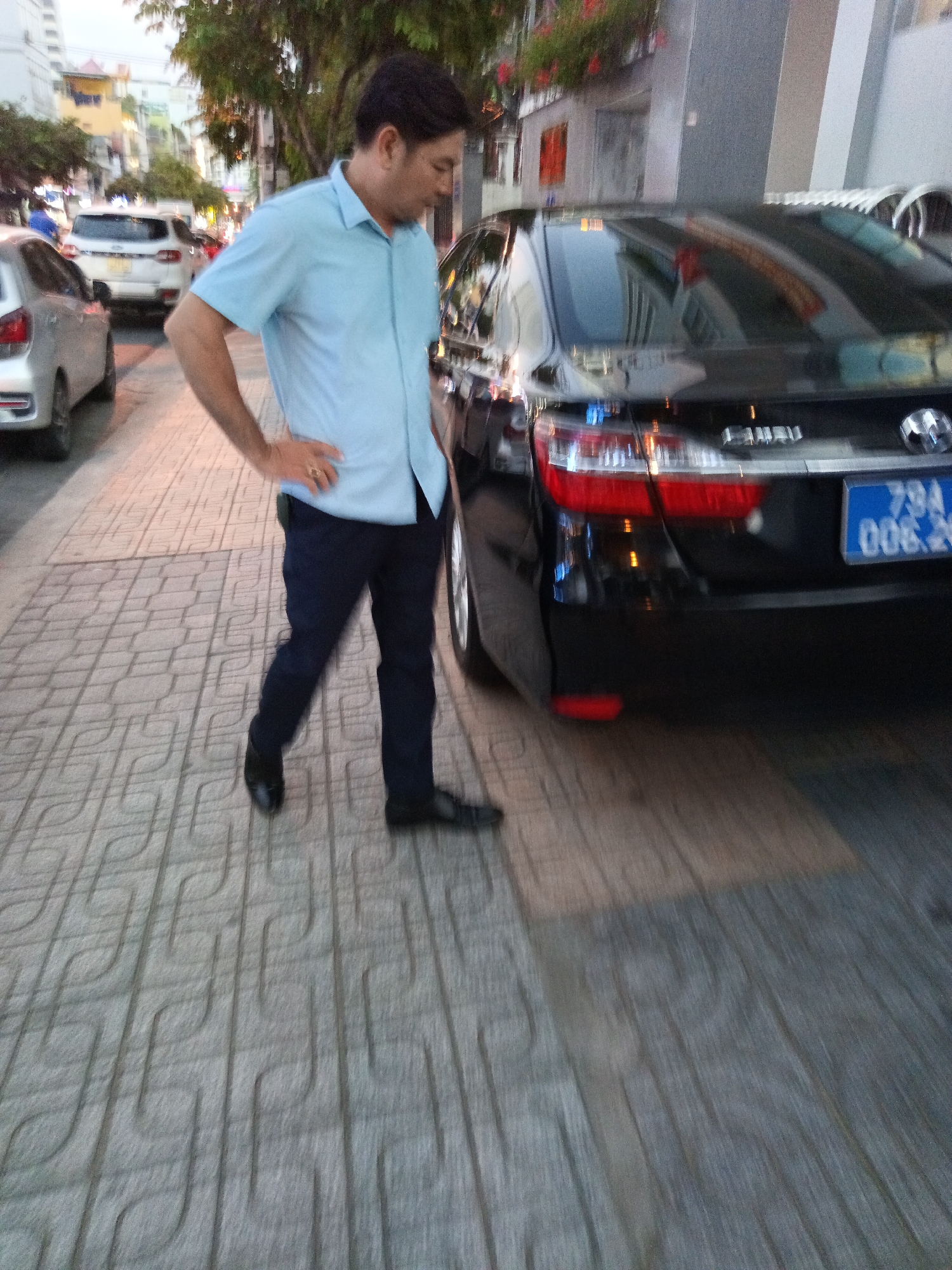 An adult male wearing a light blue collared shirt and dark pants stands on a sidewalk with a patterned tile surface. He has his right hand on his hip. Behind him, a black sedan is parked on the street. The car has a blue license plate with white lettering and numbers. In the background, other vehicles and buildings are visible, suggesting an urban street scene. The overall lighting suggests it is either dusk or an overcast day. The scene appears to be in Nha Trang, Vietnam.
