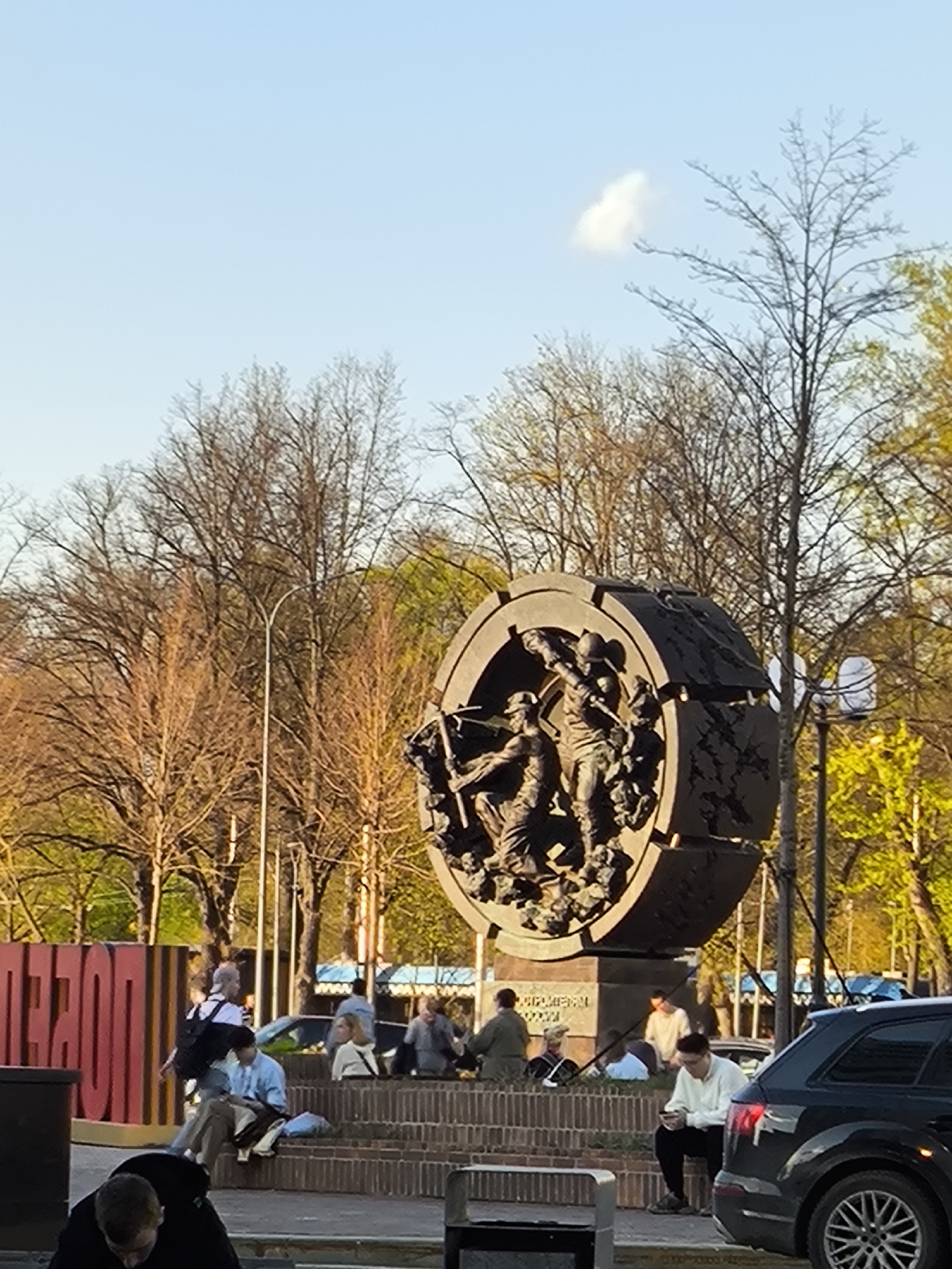 The image shows a public space in Moscow, Russia, featuring a large circular monument with a bronze bas-relief of workers. The monument sits on a brick base where several people are sitting or standing. The monument is partially obscured by trees in the background.
In the foreground, several people are casually dressed, some sitting on the brick structure and others standing nearby. A dark SUV is parked on the right side of the frame. Some large red letters are visible behind the monument, possibly spelling out "MOSCOW". The sky is partly cloudy.