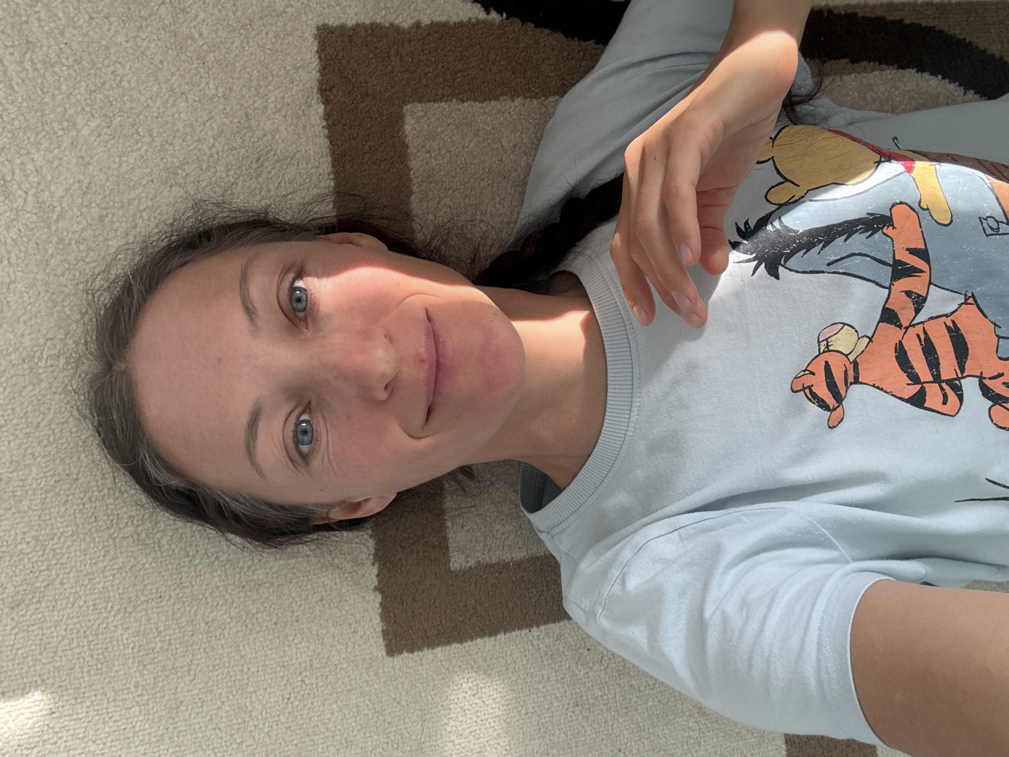A woman with braided dark hair and blue eyes is lying on a carpet with a geometric pattern. She is wearing a light blue long-sleeved shirt featuring Winnie-the-Pooh characters. The location is not identifiable as Ulyanovsk, Russia, based on the visual information.