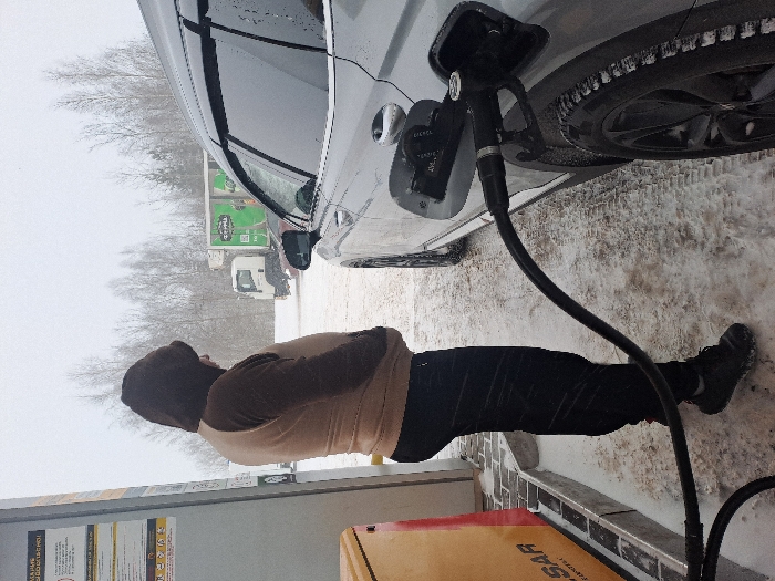 A person in a brown hooded jacket and black pants fills a gray car with gas at a snowy station. The gas pump nozzle is inserted into the car's fuel tank. Snow covers the ground, and trees and vehicles are visible in the background. The scene appears to be cold and overcast.