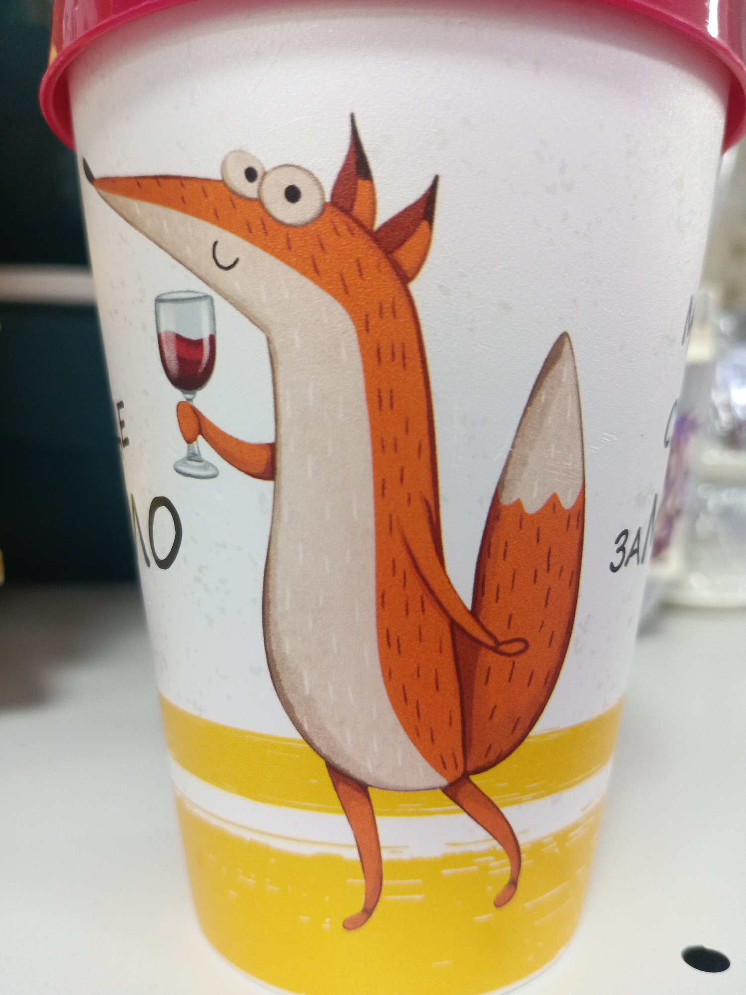 A close-up shot depicts a plastic cup with a cartoon illustration of an orange fox. The fox is standing upright, holding a glass of red wine in its paw. The cup's background is white with faint speckles, and there are horizontal yellow stripes near the bottom. The cup appears to be located in Фоки, Russia.