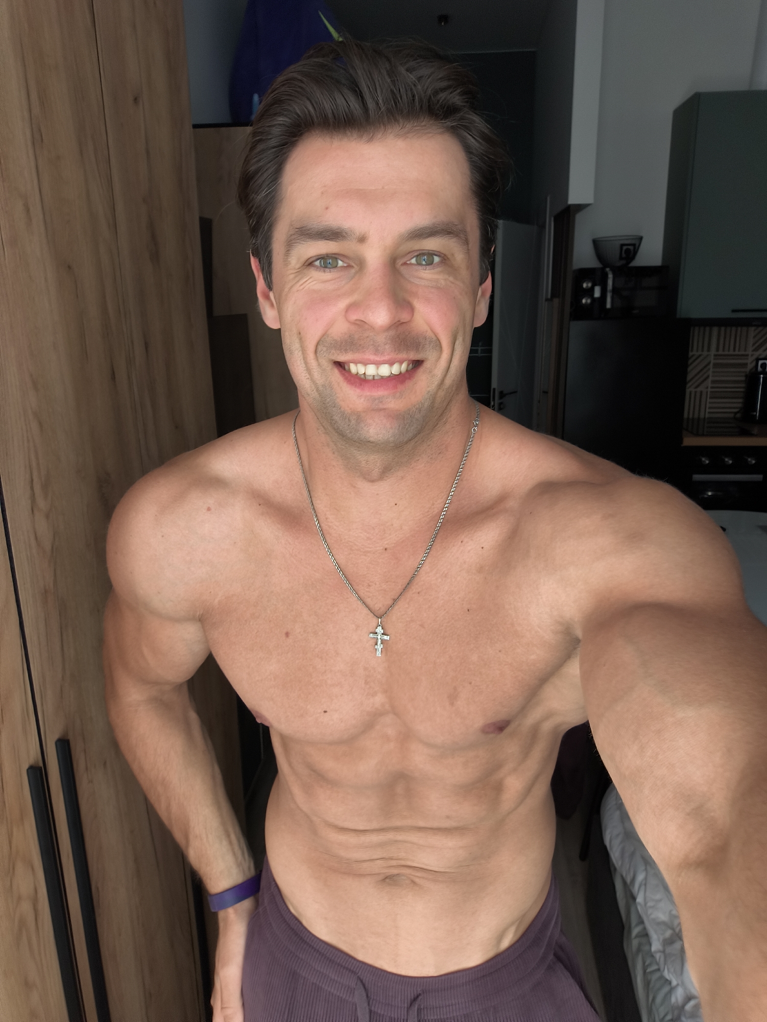 A shirtless man with a muscular physique stands facing the camera, smiling. He has short, dark brown hair and green eyes, and is wearing a silver chain with a cross pendant. The man is positioned in what appears to be a home environment, with a wooden wardrobe to his left and a kitchen area visible in the background. The overall lighting suggests it is daytime. There is no indication of the location being Moscow, Russia.