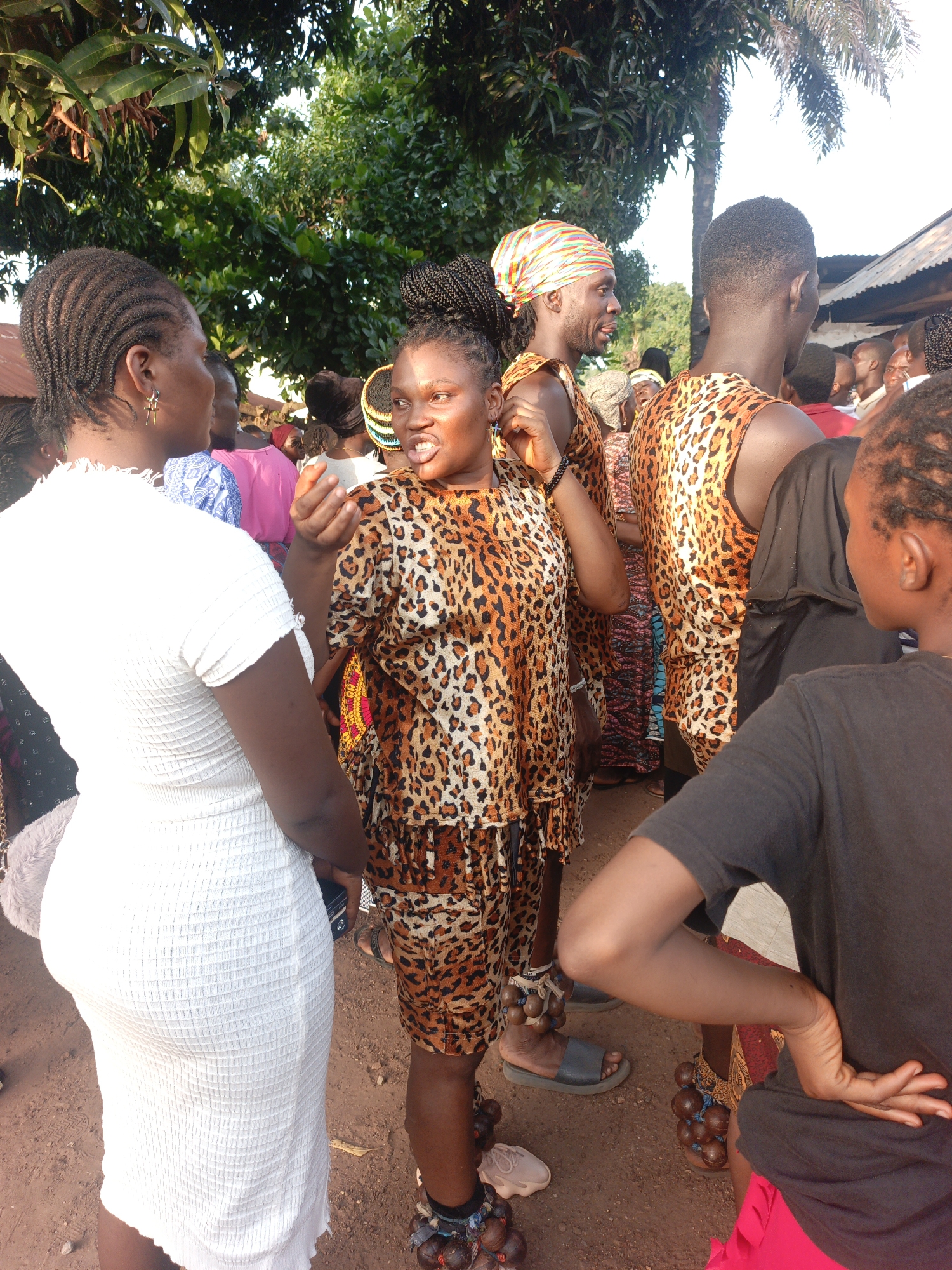 In the city of Akwanga, Nigeria, a group of people are gathered outdoors, with lush green trees forming a canopy above. The ground is a dusty, reddish-brown dirt path. In the foreground, three individuals are prominently featured. On the left, a woman wearing a white, textured, knee-length dress and a fluffy grey accessory on her left arm is seen from the waist down, facing left and engaged in conversation. In the center, a woman in a leopard-print outfit, consisting of a halter-top and a skirt, is speaking with her mouth open. Her legs are adorned with anklets made of round, brown seed pods. She is wearing dark gray flip-flops. To the right of her, a person wearing a black t-shirt and bright pink shorts is standing with their back to the camera, with their right hand resting on their hip. Their left arm is bent, with their hand extended forward. Further in the background, several other individuals are visible, some wearing similar leopard-print clothing, including a man with a colorful headscarf, suggesting a cultural event or celebration. The lighting indicates it is daytime, with sunlight filtering through the trees.