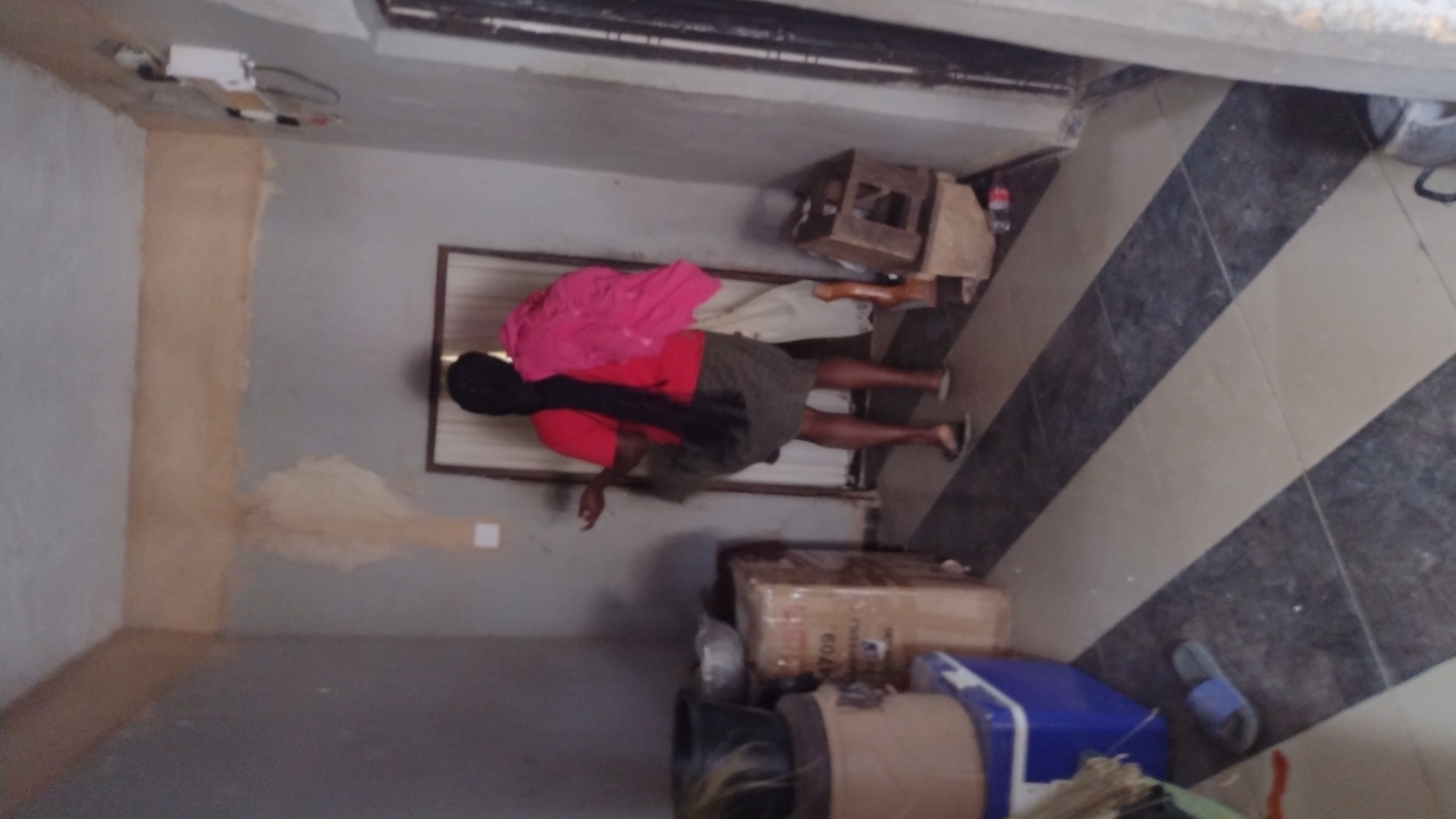 A person with long black hair is lying on the floor in a room. The person is wearing a red shirt and a dark green skirt. A pink garment is bundled on their back. There is a wooden stool, a wooden box, cardboard boxes and a blue container nearby. The room has mottled gray and light tan walls and a tile floor with a diagonal pattern of dark gray and light tan tiles. The location is potentially within Jos, Nigeria.