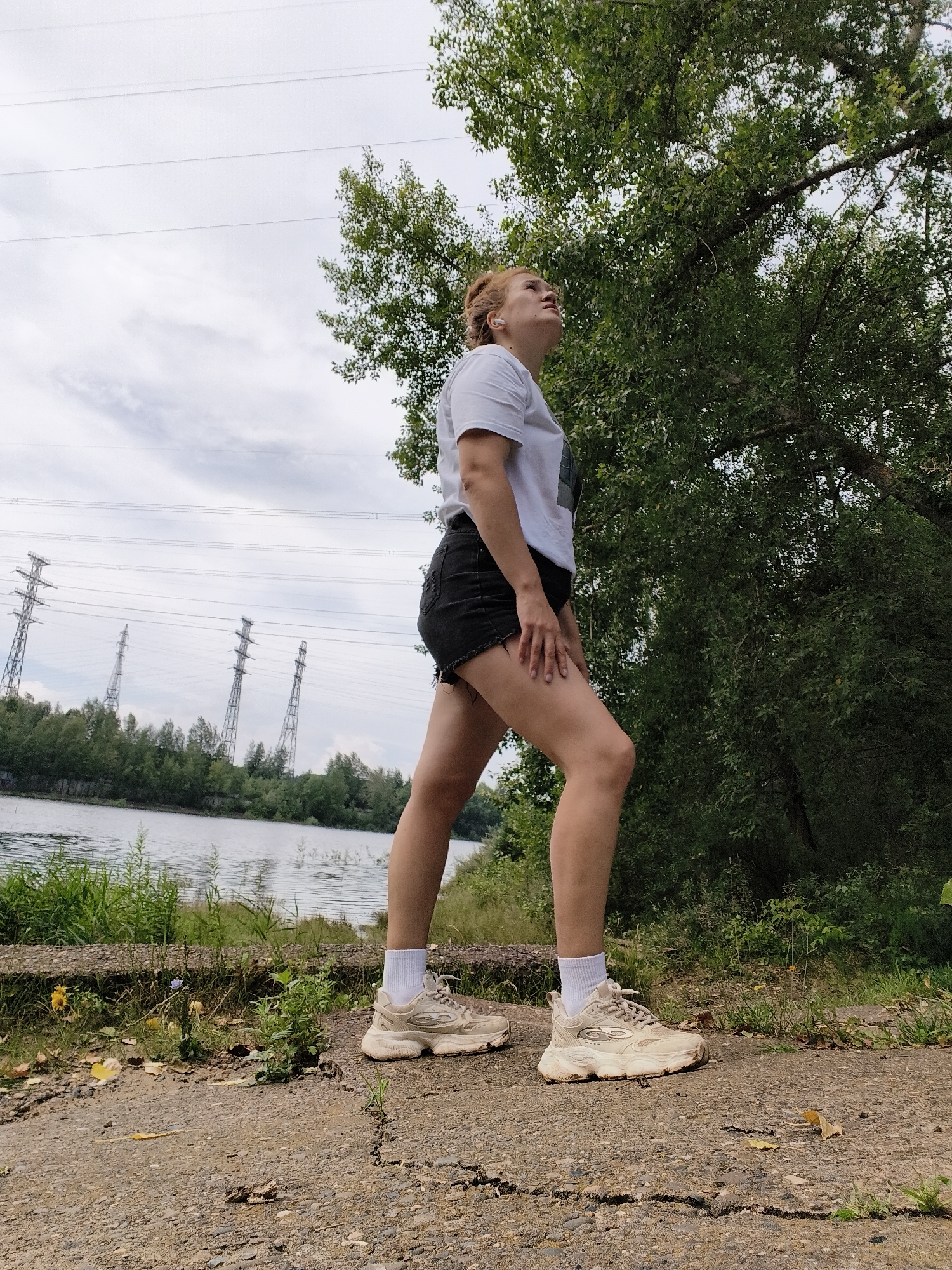In an outdoor scene under a cloudy sky, a young woman with reddish-blonde hair pulled back stands facing left, looking upwards. She wears a white t-shirt, black denim shorts, and beige athletic shoes with white socks. The woman is positioned on a cracked, gravelly path. To her left, a body of water is visible, bordered by lush green grass and vegetation. In the background, across the water, a line of trees separates the water from a row of tall, industrial power line towers. A large tree with dense green foliage is to the right of the woman, partially obscuring the sky.
