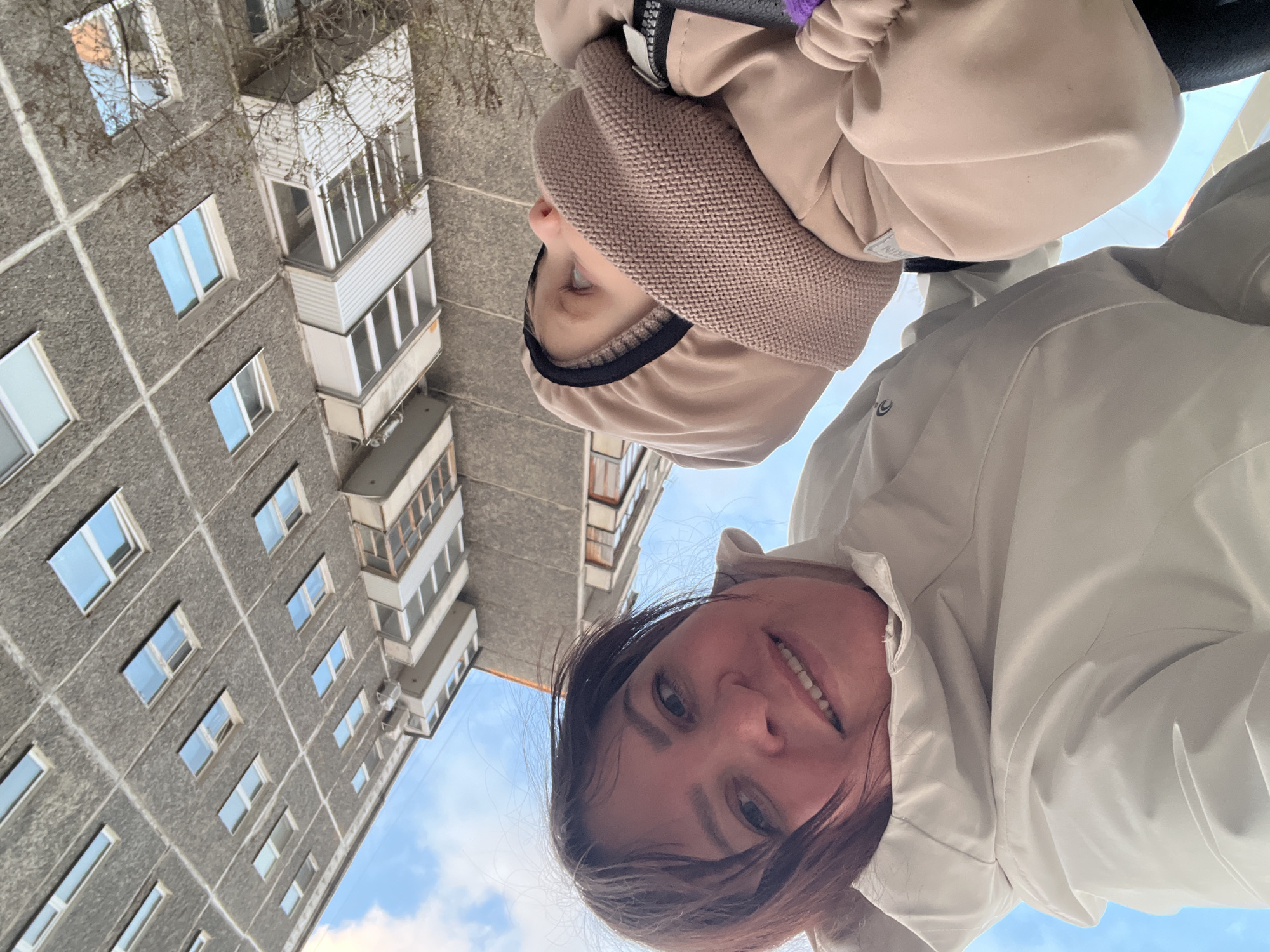 A selfie captures a woman and a child from a low angle, with a Soviet-era apartment building looming in the background. The woman, positioned closer to the camera, smiles with her dark hair visible beneath a light-colored jacket. The child, wearing a tan hooded jacket and knitted hat, stands behind her. The multi-story building is constructed from grey concrete panels with repetitive window placement, indicating a typical residential structure from the Soviet period. The blue sky is partially visible. The image was taken in Perm, Russia.