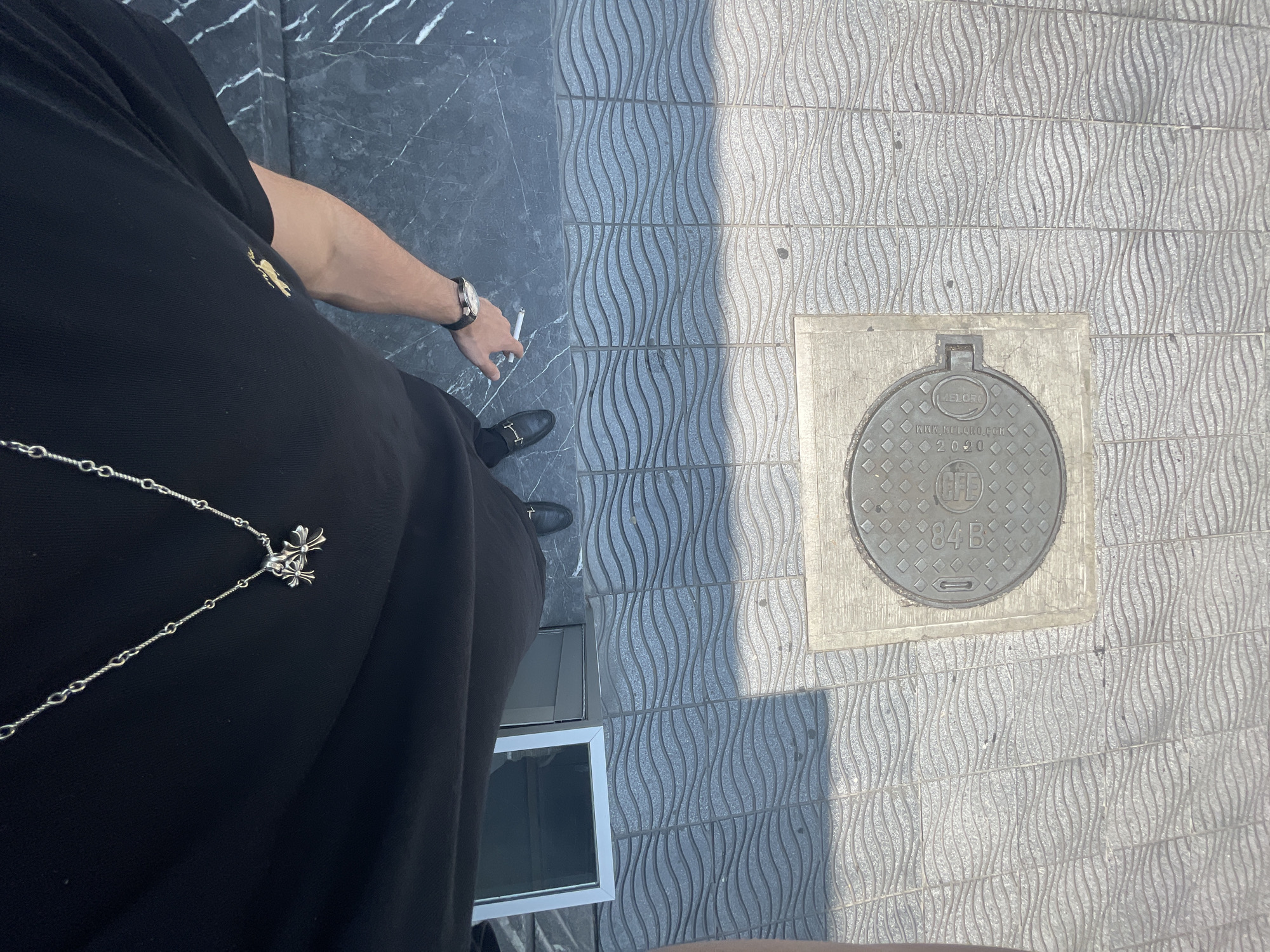 The photograph is taken from an overhead perspective. A person wearing a black polo shirt, a silver chain with cross pendants, and a watch is visible. They are holding a cigarette. The scene includes pavement with a textured, wavy pattern, a marble-like surface, and a manhole cover labeled "ME LOBO" and "2020". Based on the manhole cover inscription, the location is in Mexico City, Mexico. A window of an unknown building is also visible.