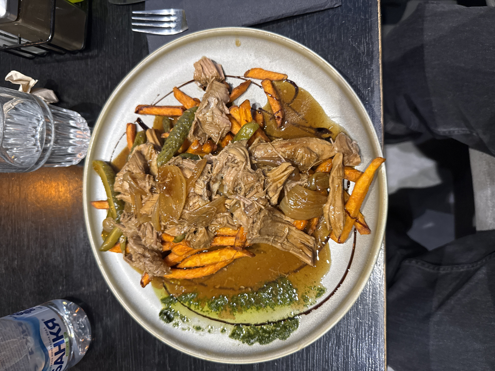 A top-down shot depicts a plate of food with shredded meat, sweet potato fries, and pickles covered in gravy and a green sauce. The plate rests on a dark gray table next to a fork, a water bottle, a glass, and what appears to be a metal container. A person's lap is visible on the right. The location is unspecified, but the city is given as Blagoevgrad, Bulgaria.