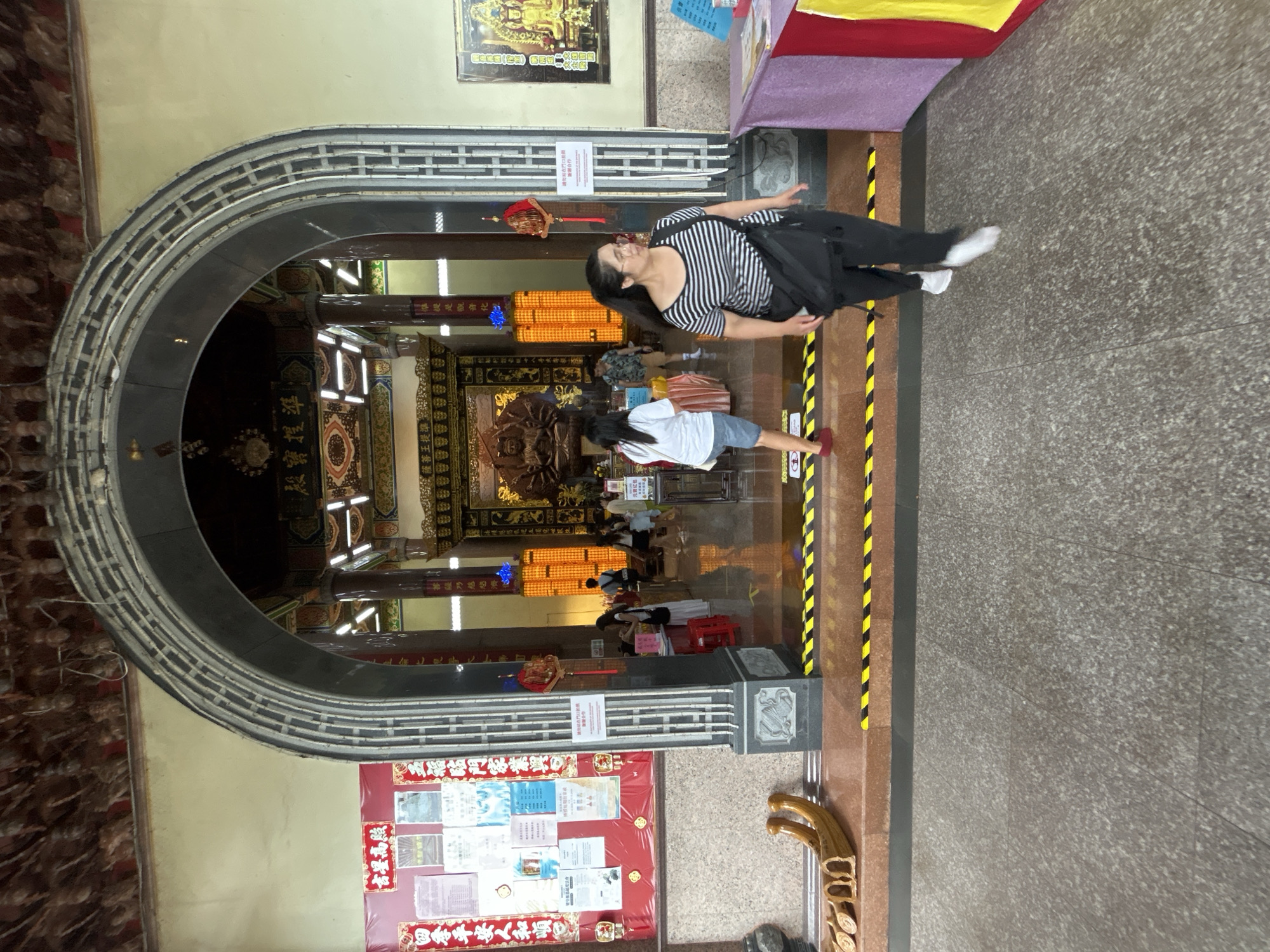 In George Town, Malaysia, a woman with dark hair, wearing glasses, a black and white striped t-shirt, and black pants, walks towards the foreground of the image. She carries a black bag over her shoulder. Behind her, a temple interior is visible through an arched entryway. Inside the temple, several people are present, including a woman in a white t-shirt and denim shorts walking away from the camera. The temple features ornate decorations, including a large golden statue and illuminated pillars. The floor in the foreground is a textured grey stone, with yellow and black caution tape marking a boundary on a step leading into the temple. To the left of the entrance, a bulletin board with various papers is displayed on a red background. To the right, a table covered with a purple cloth displays some items. The architecture of the entrance features decorative grey stone elements.