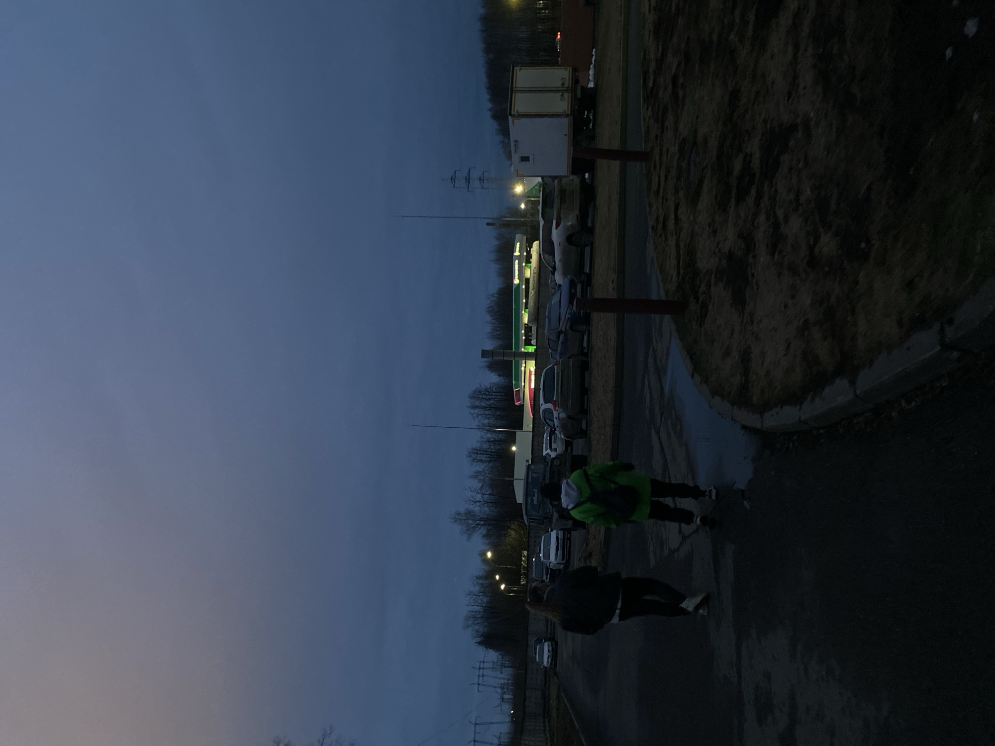 Two people are walking on a wet asphalt surface alongside a roadside area with parked cars, a gas station, and green vegetation. The sky is darkening, indicating dusk or twilight. The location appears to be in or near a town or city, evidenced by streetlights and power lines. Given the request for a location in Izhevsk, Russia, the gas station brand and style of architecture is consistent with locations in that region.