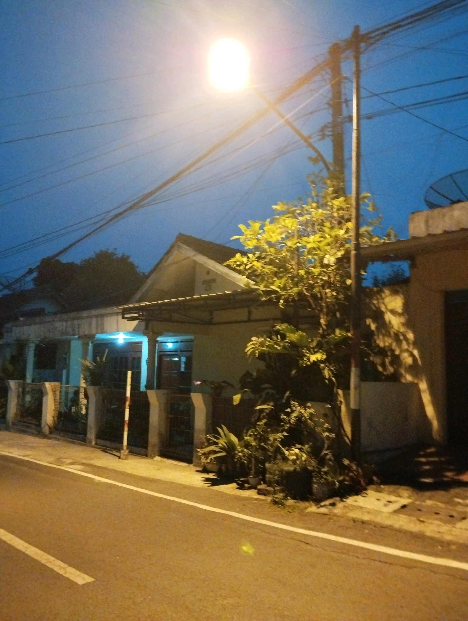 The image depicts a street scene at dusk or night in Salatiga, Indonesia. A street lamp illuminates the foreground, casting a bright glow on the pavement and a nearby tree. A pole supporting overhead electrical wires stands prominently in the center of the frame. To the right, a building with a white section and a satellite dish is visible. In the background, a residential building with an extended porch is visible, lit by two blue lights. The road is paved and features white lane markings. The sky is a deep blue, indicative of twilight. The overall impression is one of a quiet residential street illuminated by artificial light.